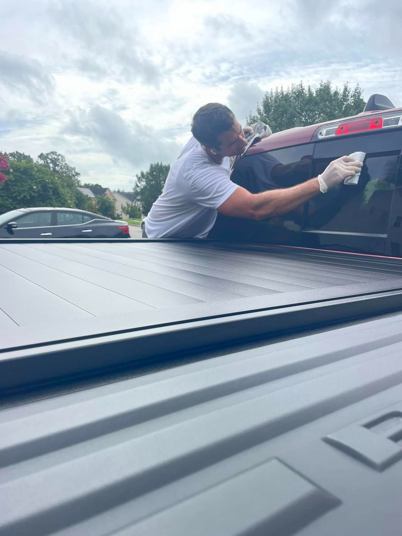 Person wearing gloves cleaning a black truck bed. The truck is parked outside on a cloudy day.