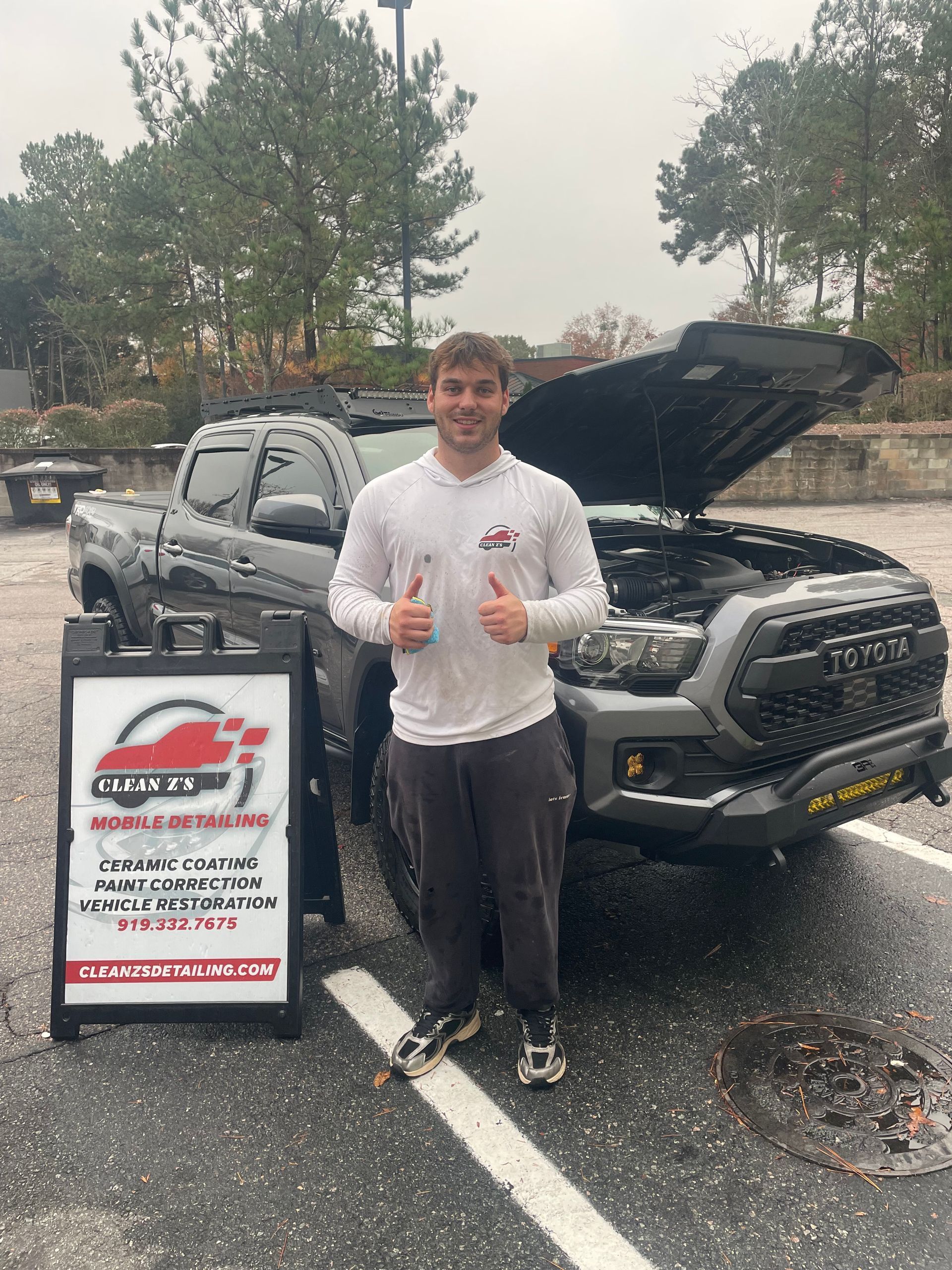 Man giving thumbs up next to a black Toyota Tacoma with its hood open, roadside sign.