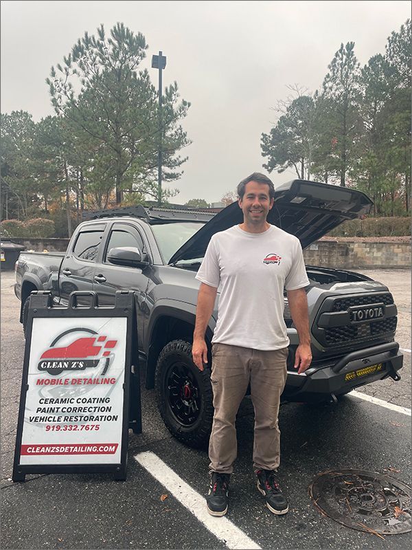 Man stands by a truck with an open hood, an advertising sign in front. The truck is dark gray with black rims.