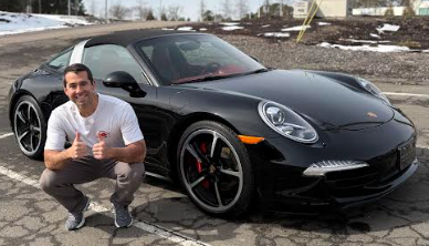Man kneels beside black Porsche Targa with thumbs up, parked in a lot with snow on the side.