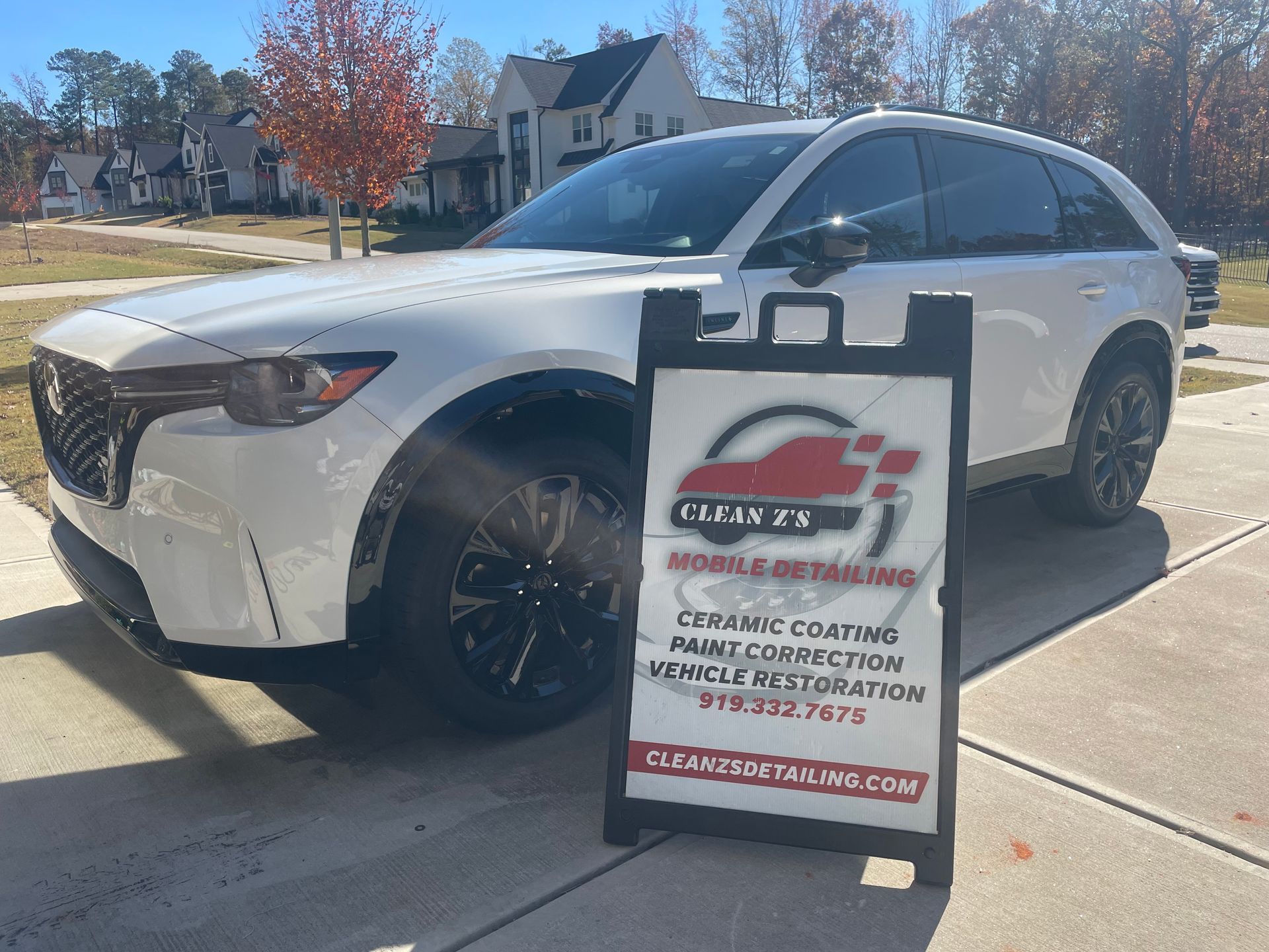 White Mazda CX-9 SUV parked on a driveway with an advertising sign for mobile detailing services.