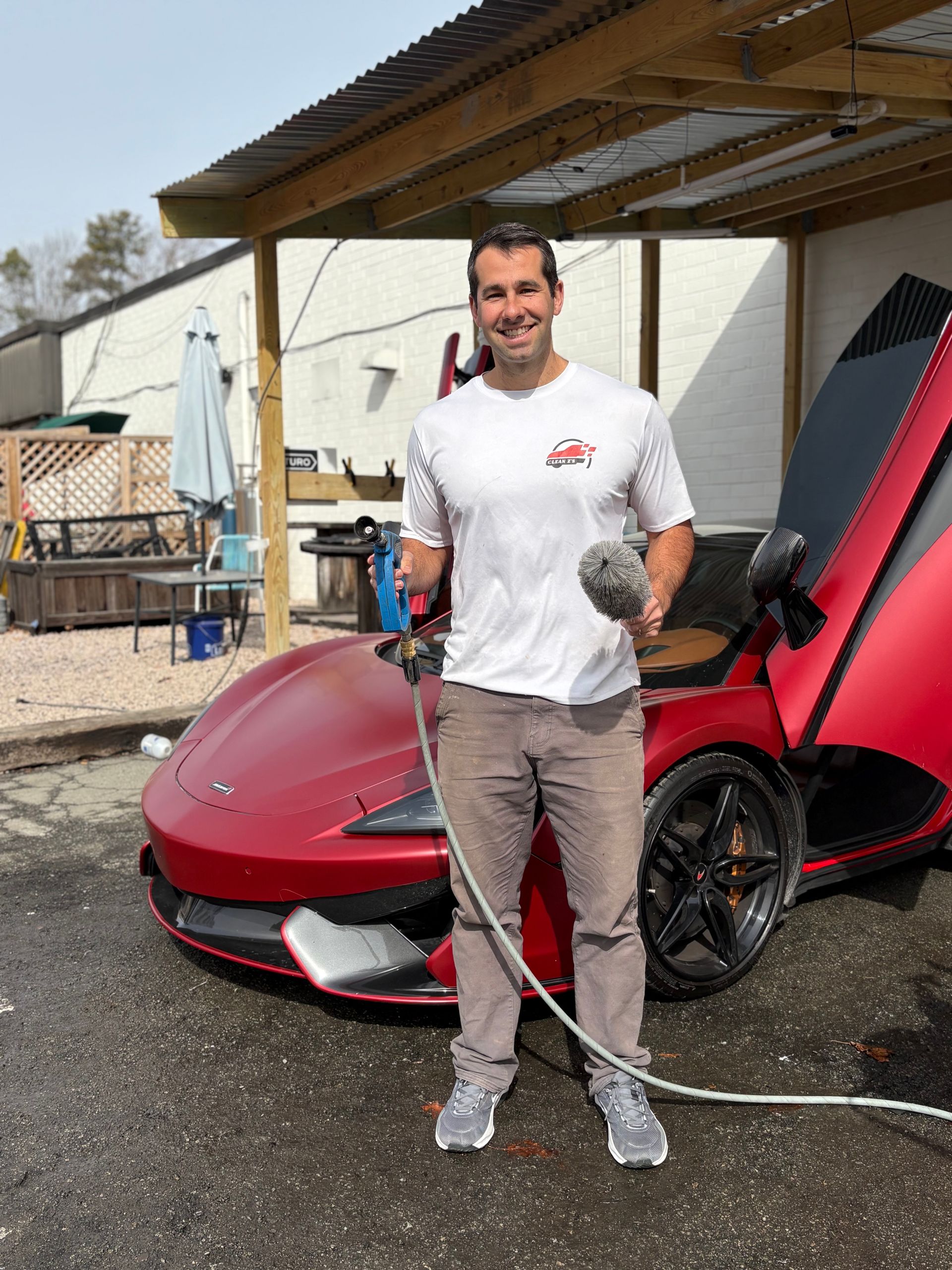 Man washing a red sports car. He stands smiling, holding a hose and sponge, with open doors.