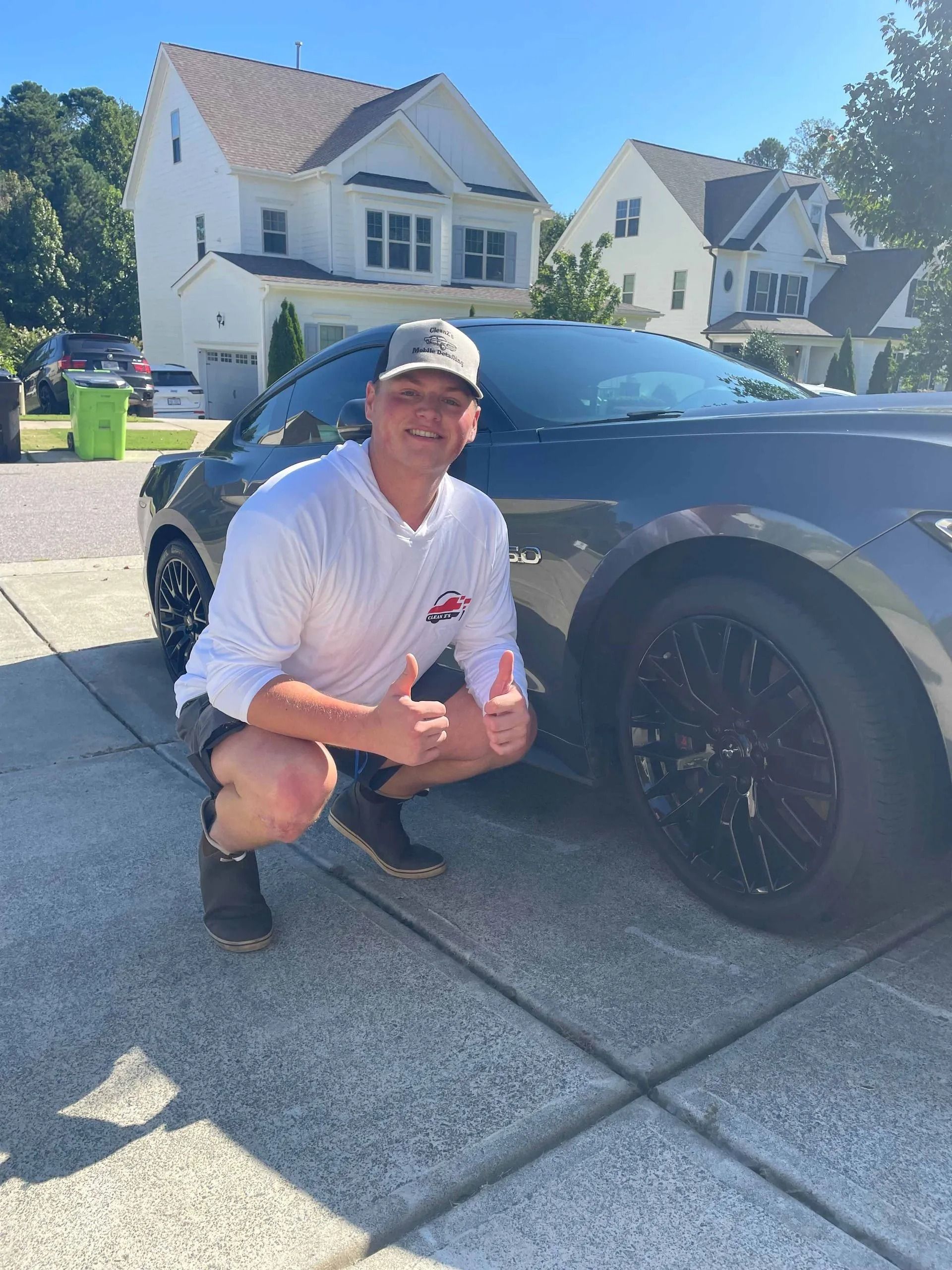 Man in a hat squats, giving thumbs up next to a dark gray car on a driveway in front of a house.