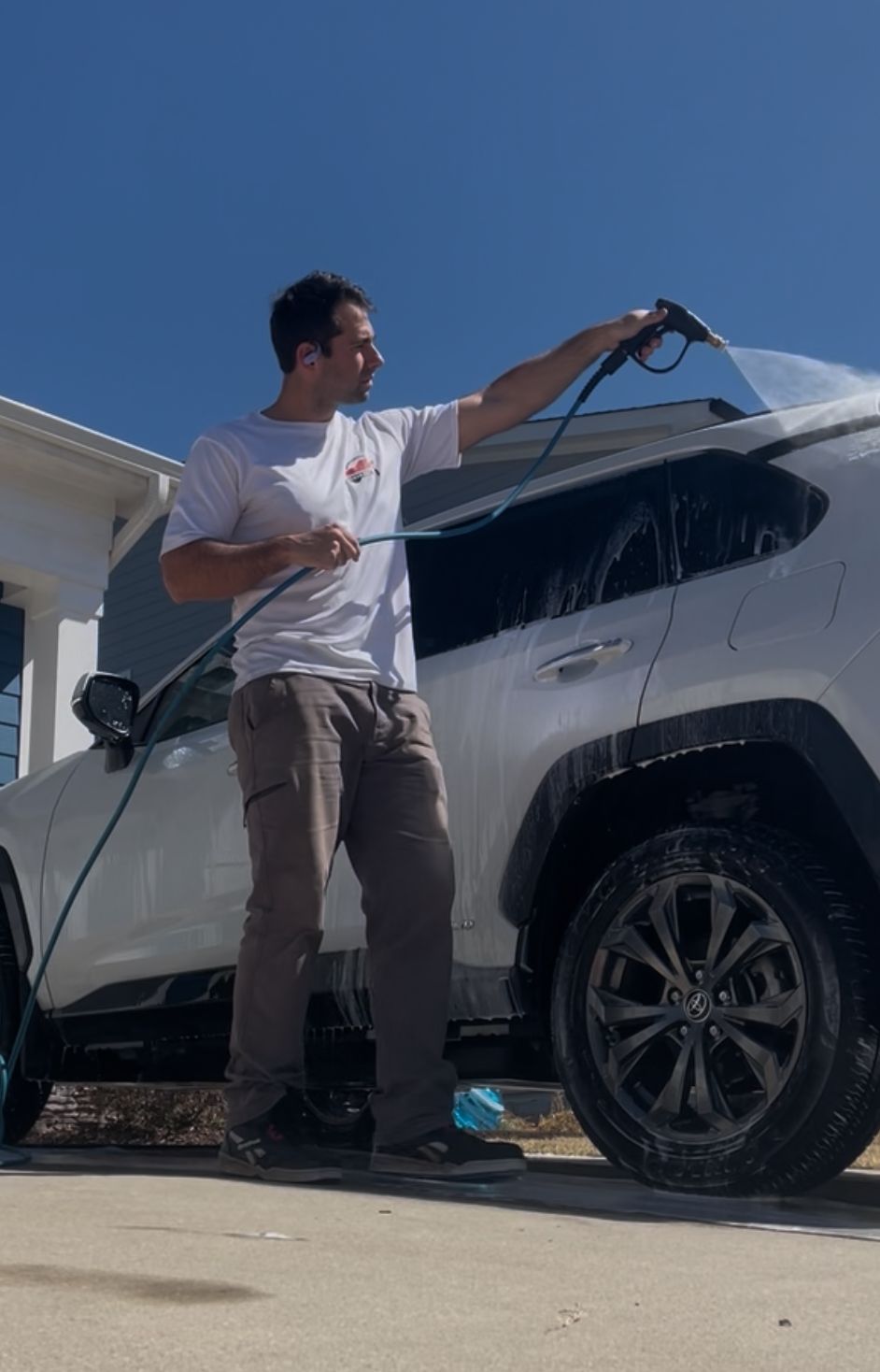 Man washing a white SUV with a hose on a sunny day.