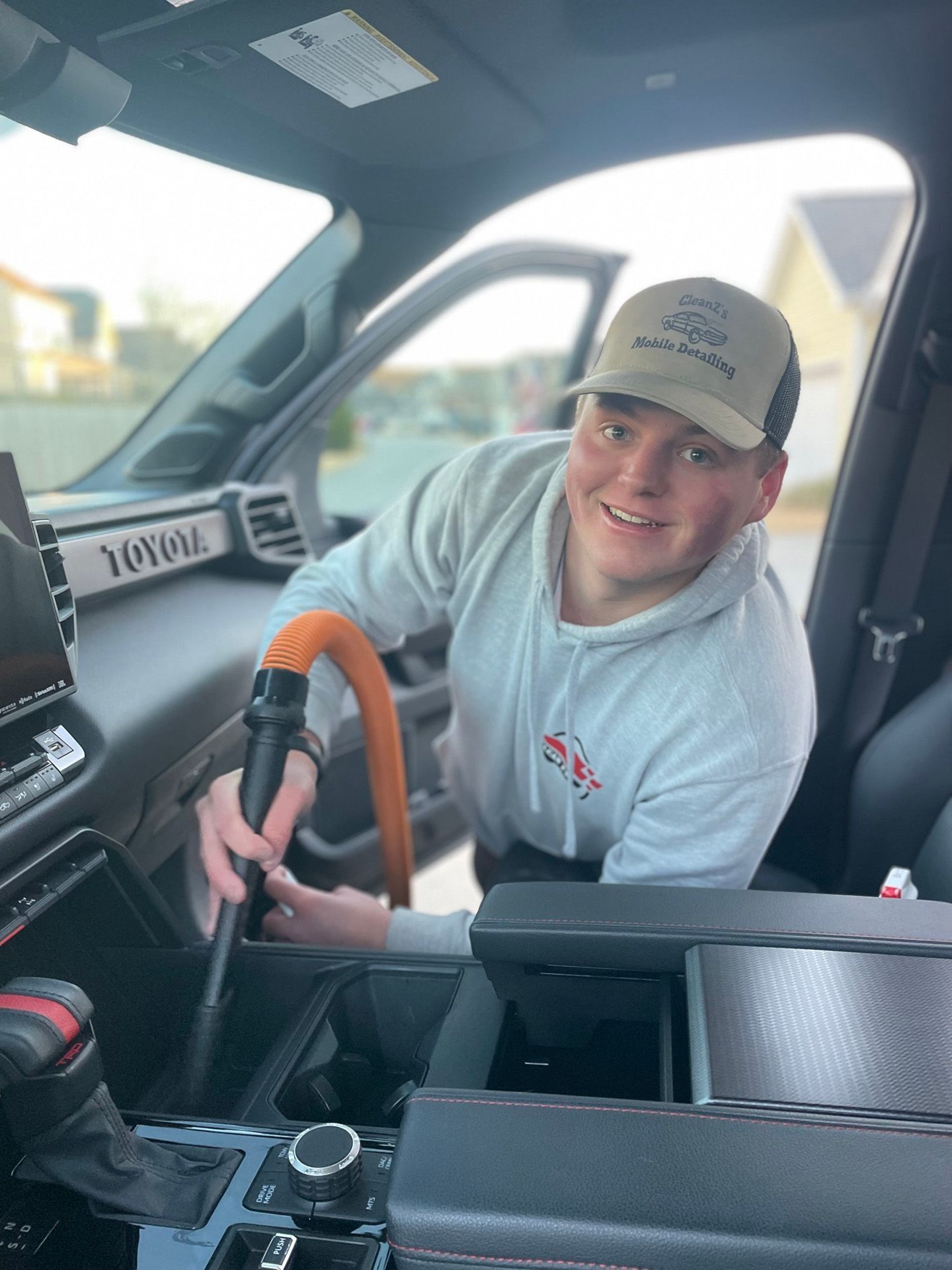 Man vacuuming the interior of a Toyota truck, smiling. He wears a hat and a gray hoodie.