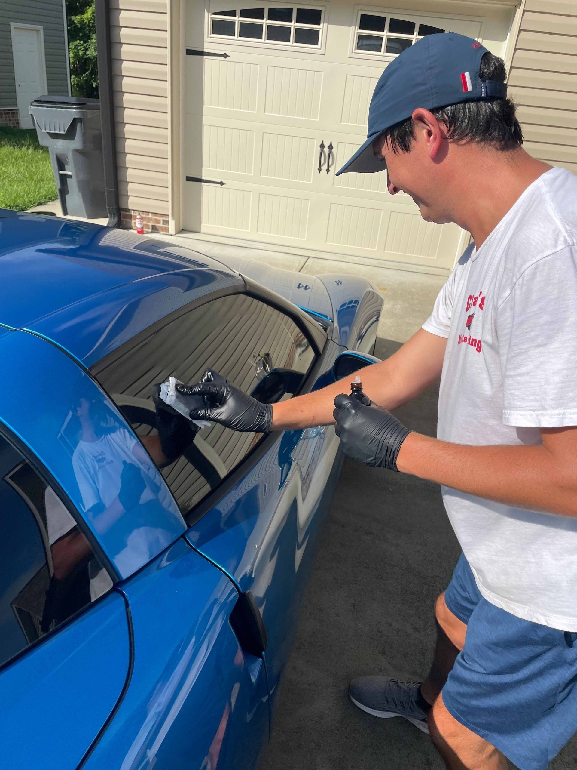 Man in a blue hat and shorts cleaning a blue sports car window outside a garage.