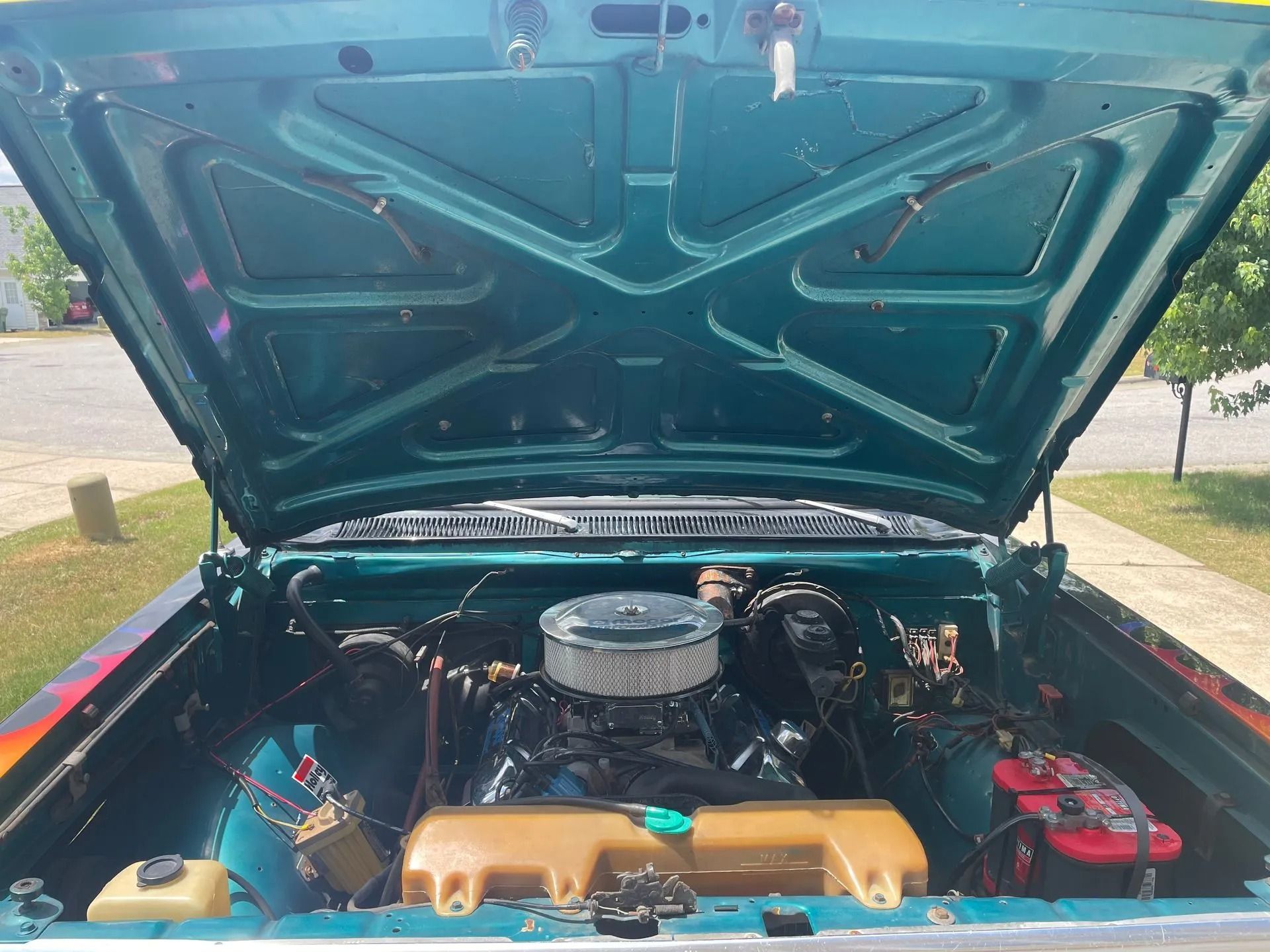 Open turquoise car hood reveals engine bay with engine and battery.