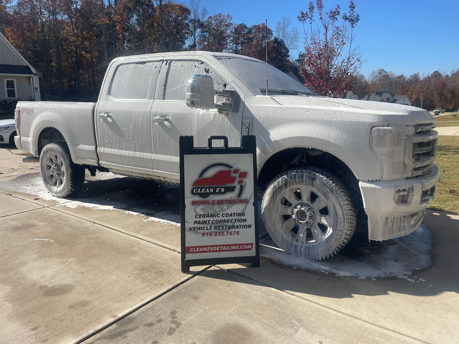 White truck covered in soap suds at a car wash, with a sign advertising car detailing services.