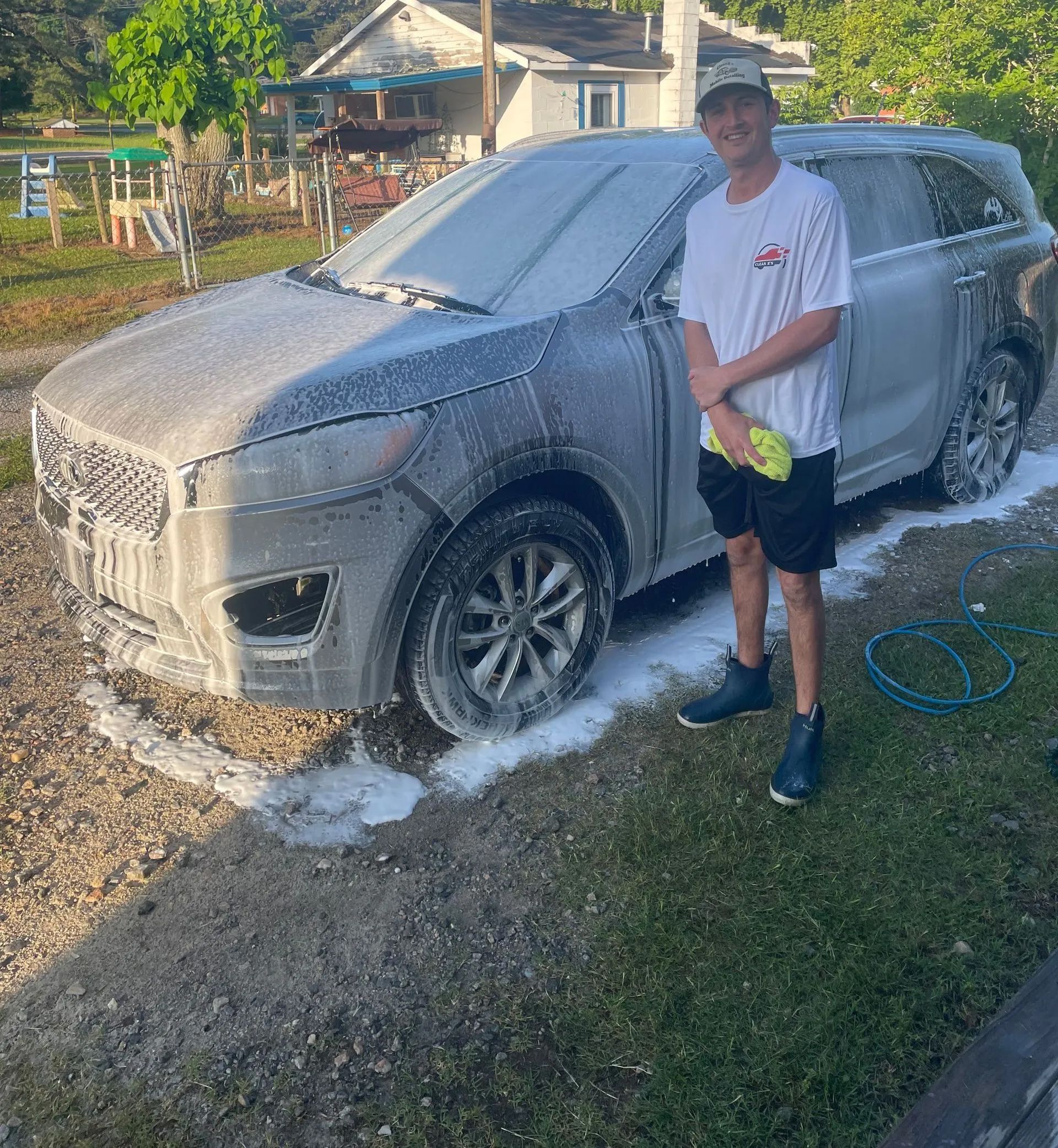 Man washing a gray car with soapy foam, standing in front of a house.