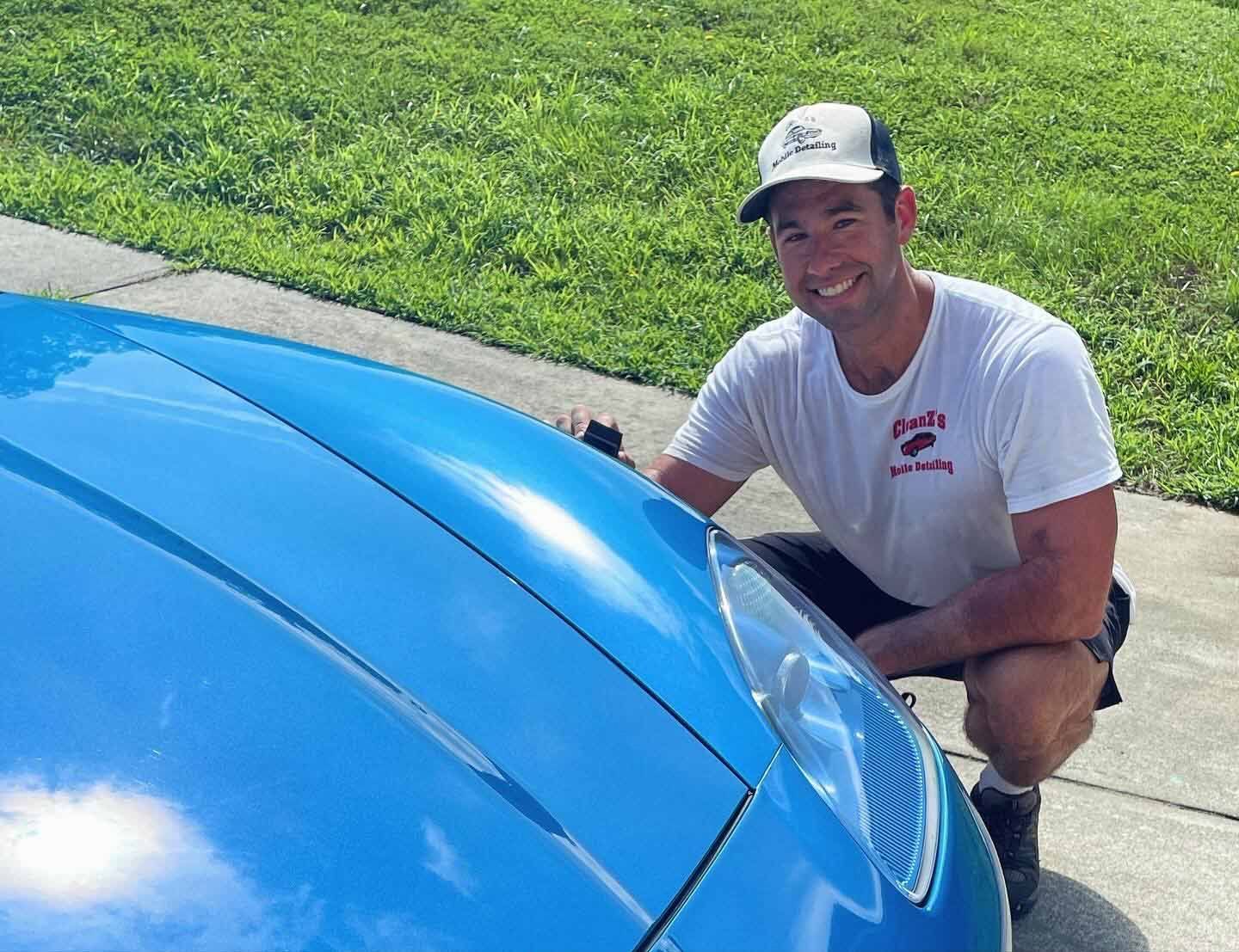 Man in cap and t-shirt squats next to a bright blue car, smiling outdoors.
