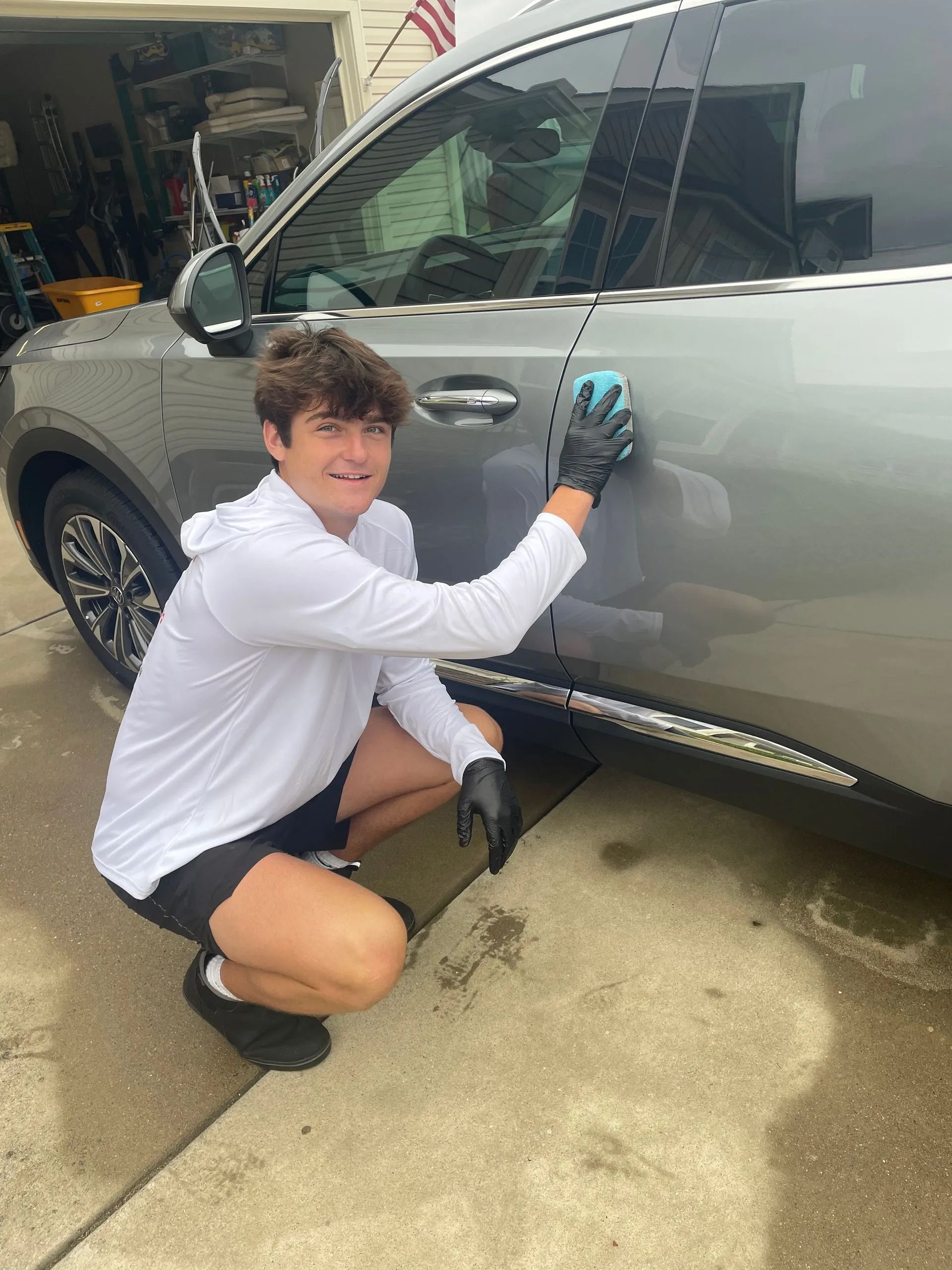 Teenager washing a silver SUV; smiling, kneeling, wearing gloves, shorts, and a white long-sleeve shirt in a driveway.