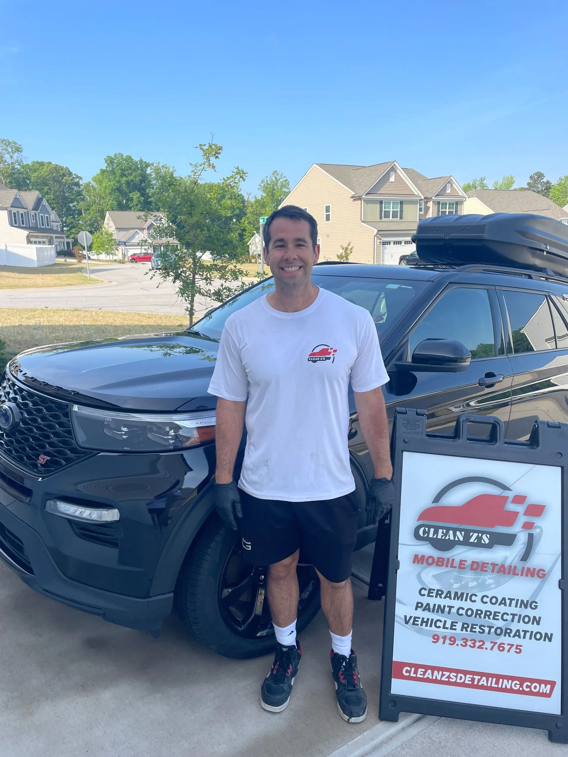 Man in front of black SUV with sign for automotive restoration. Outside, sunny day.