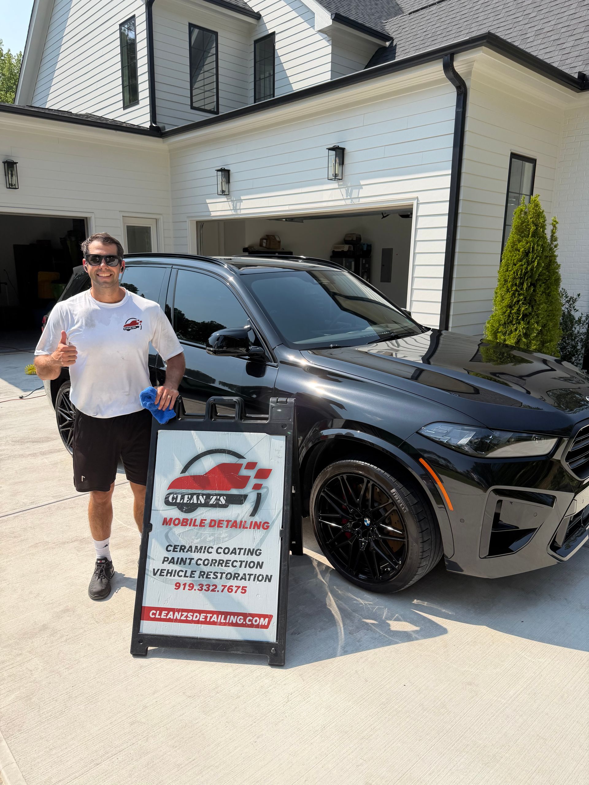 Man standing beside a black SUV and dealership sign in a driveway in front of a house