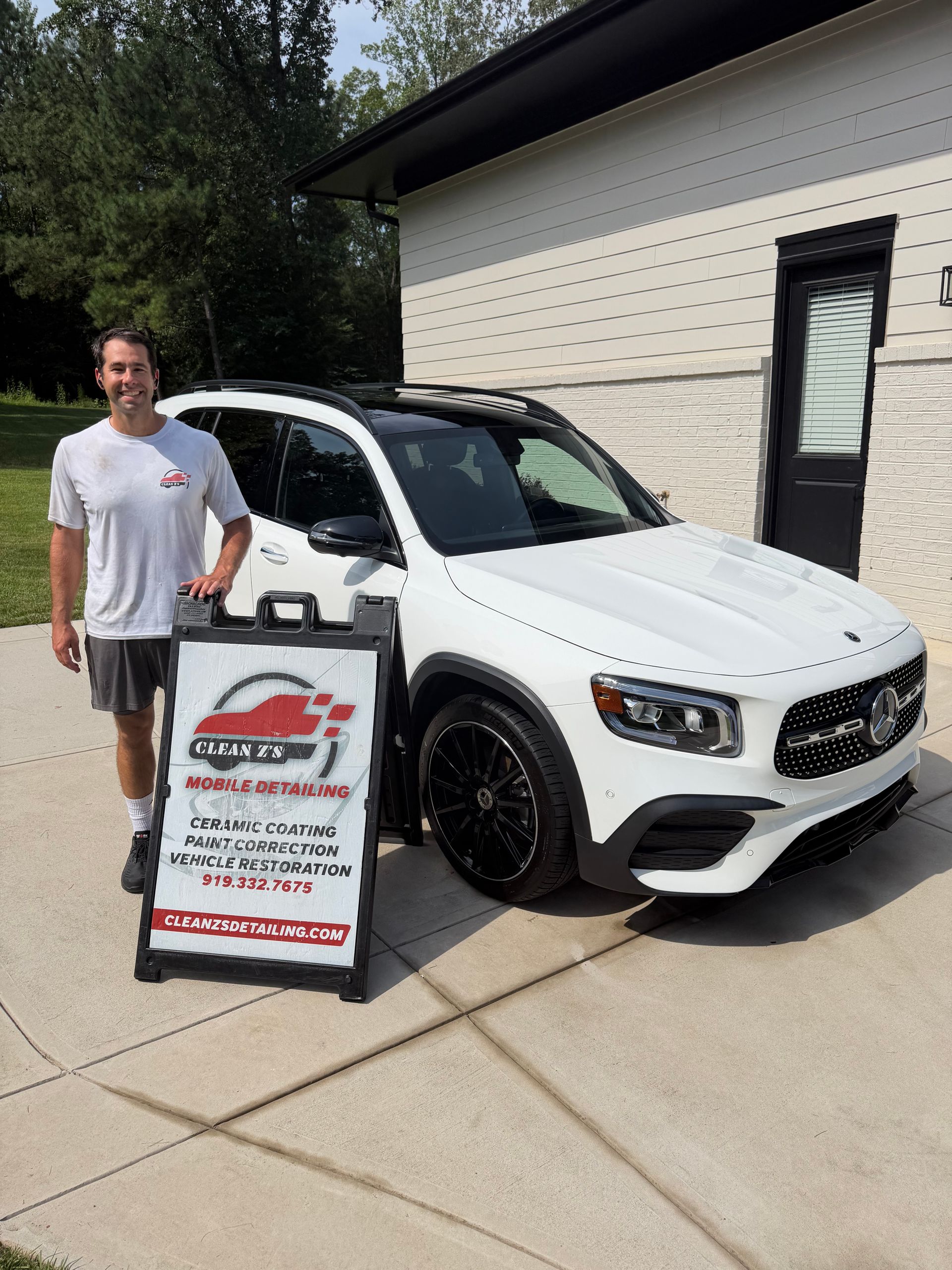 Man standing beside a white SUV and a dealership sign in a driveway.