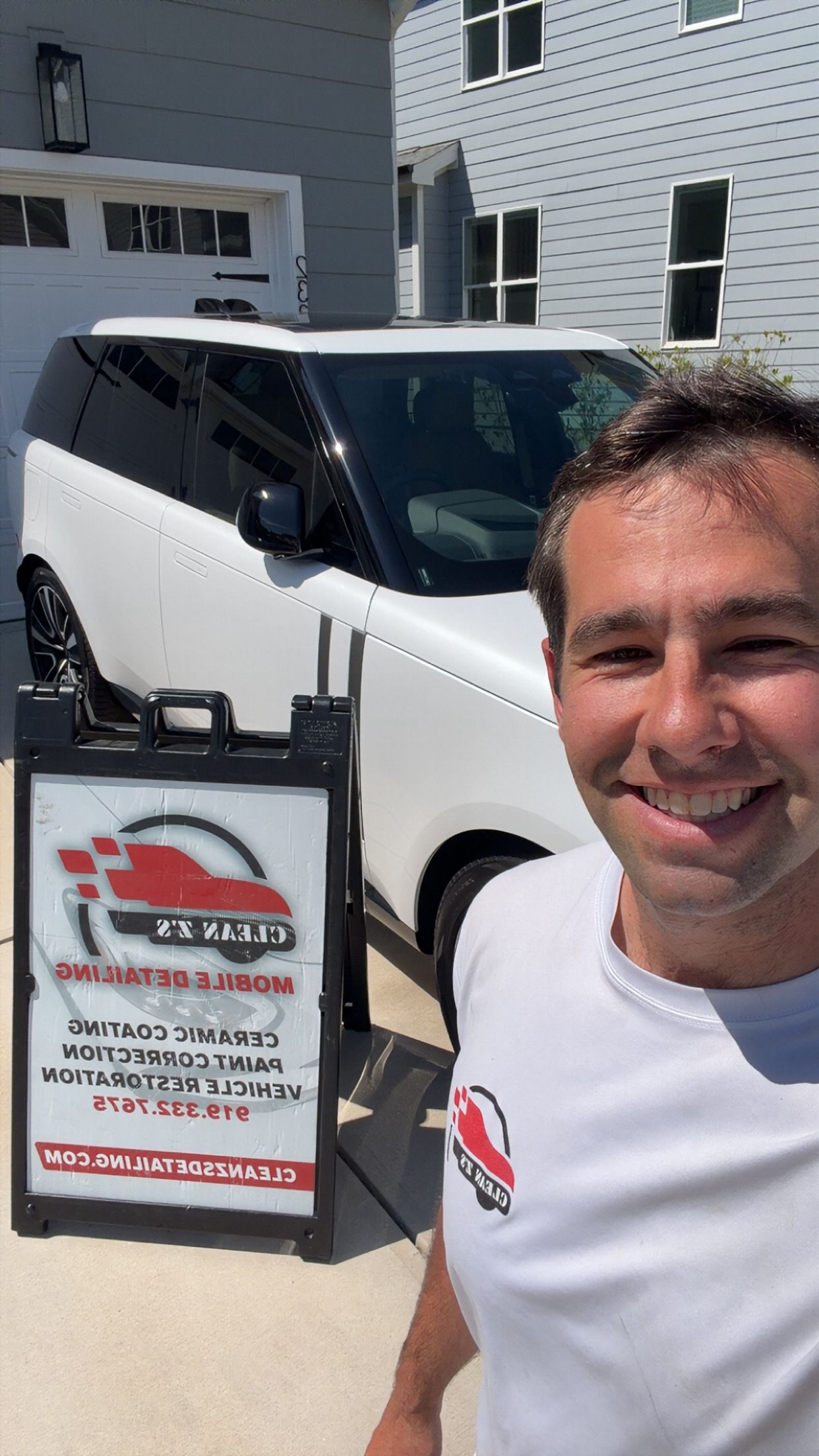Man smiling beside a white van with open side door and a sign, parked in front of a house.