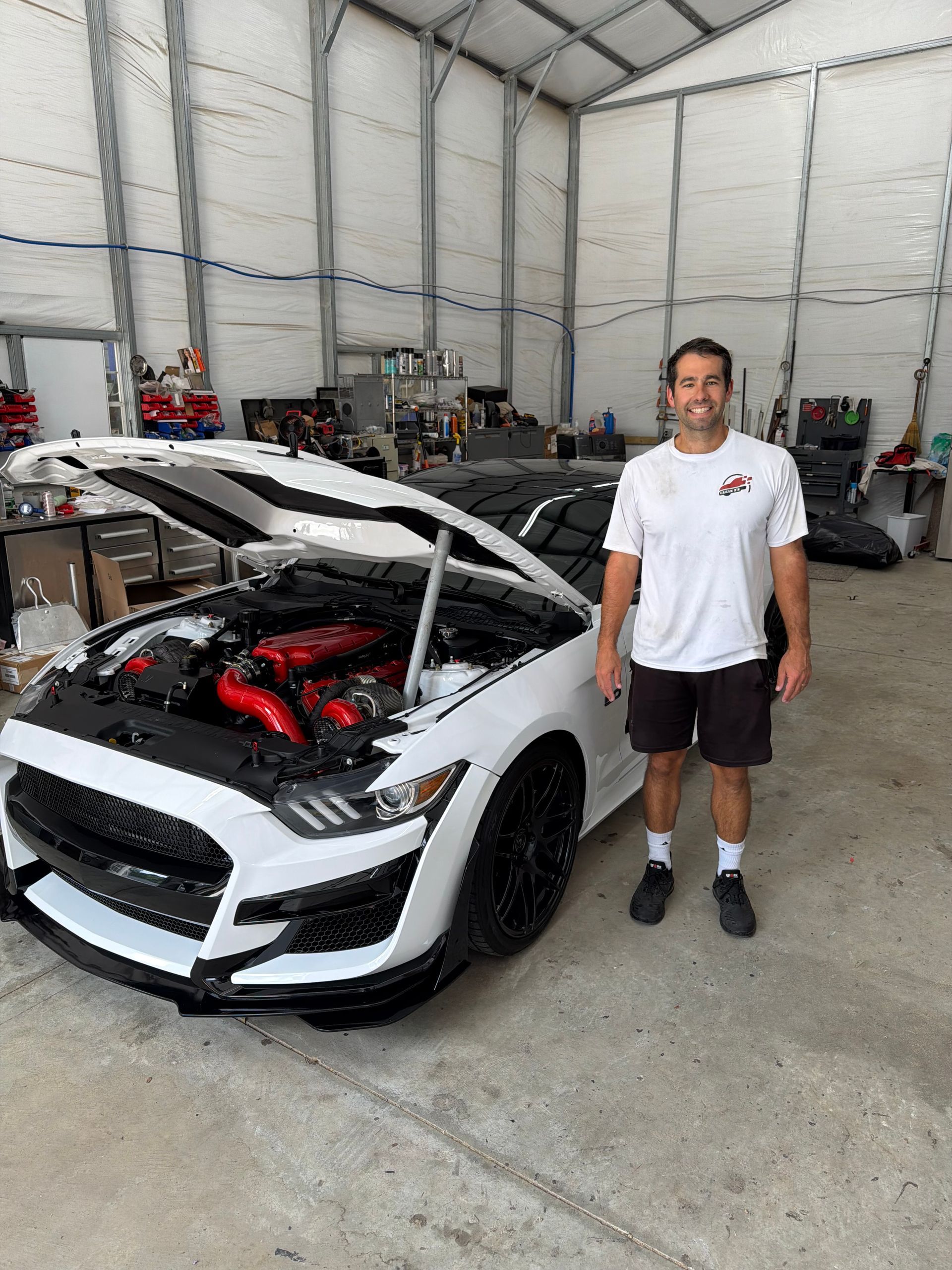 Man standing beside a white sports car with its hood open in a garage, showing a red engine.