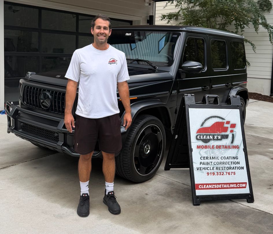 Man standing beside a black SUV in a driveway next to a detail service sign.