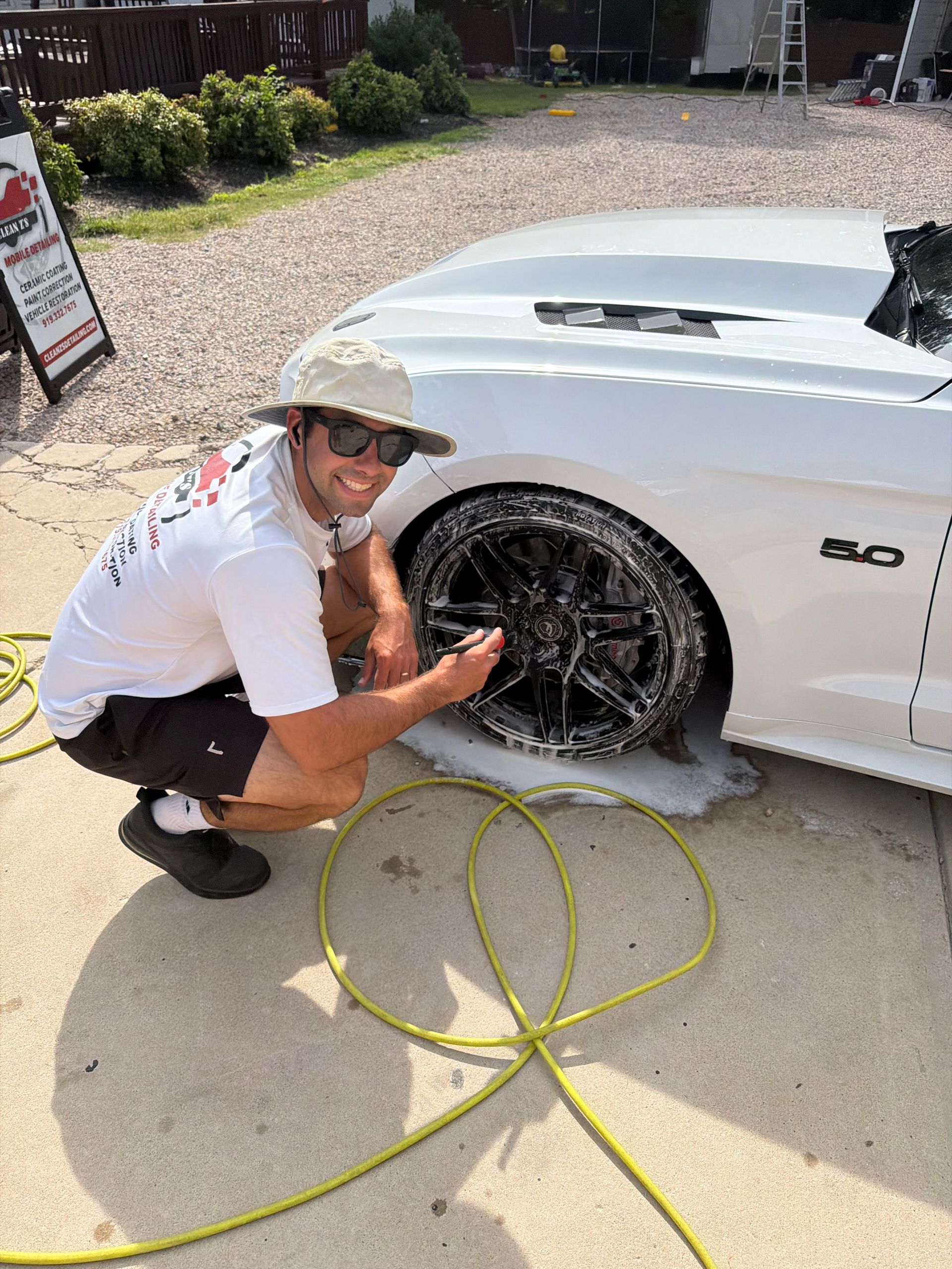Man washing a white car’s wheel with soapy water and hose in a driveway