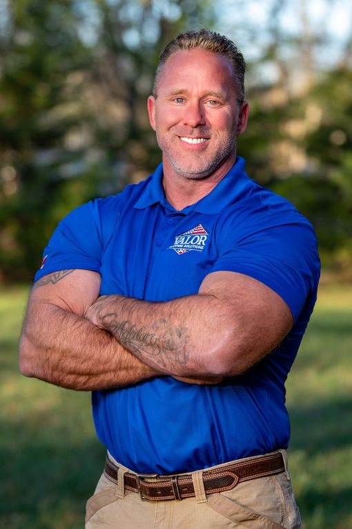 Man with crossed arms, smiling, wearing blue polo shirt and khaki pants outdoors.