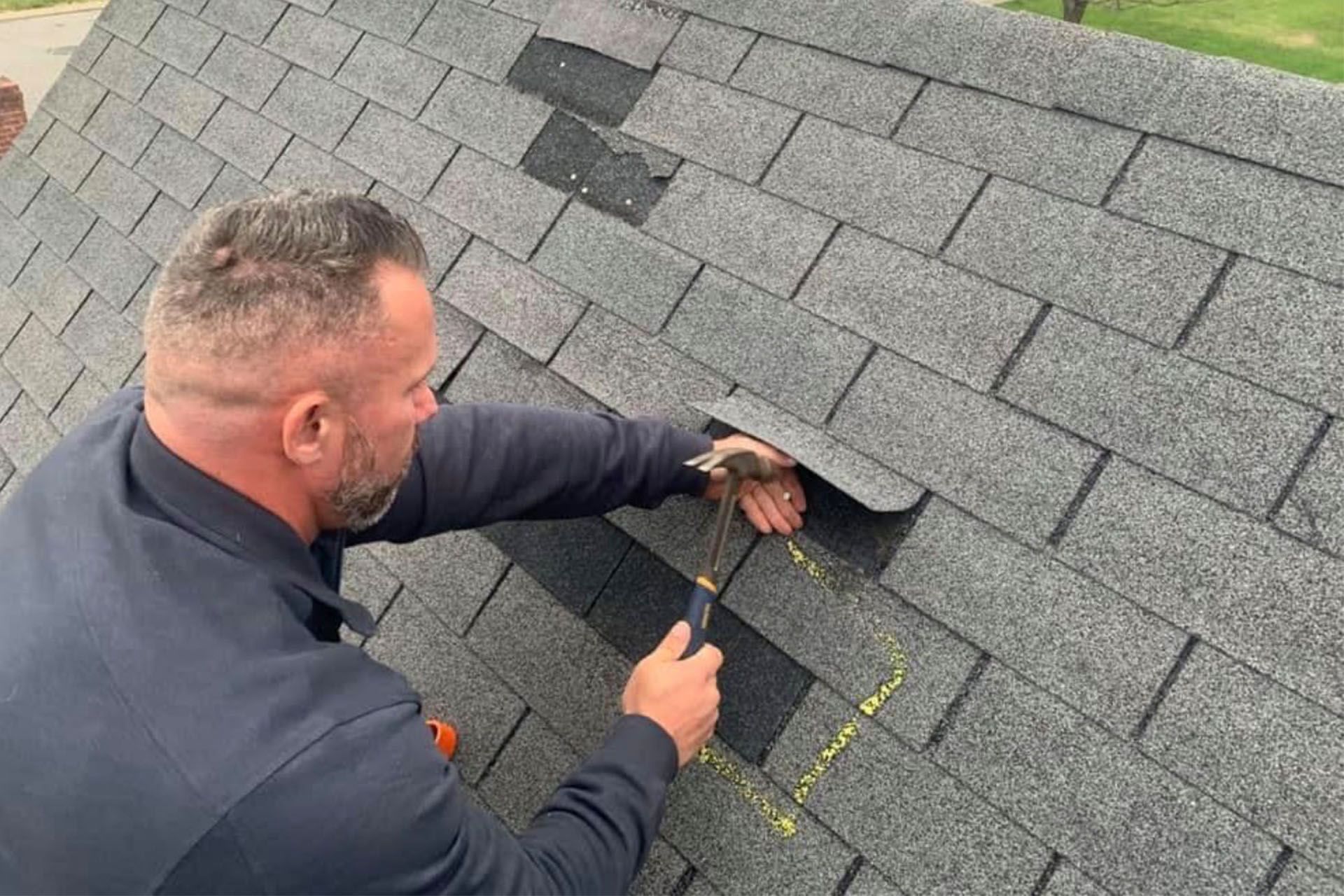 Man repairing roof shingles with a hammer; gray asphalt shingles, outdoors.