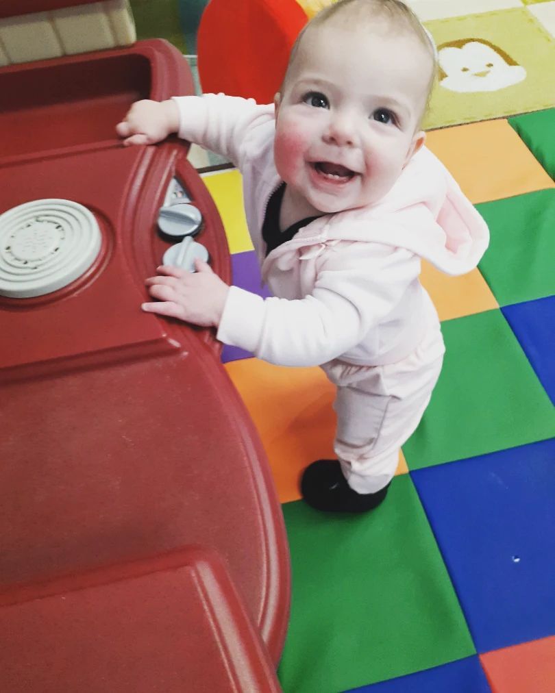 A baby in a pink outfit is standing on a colorful mat