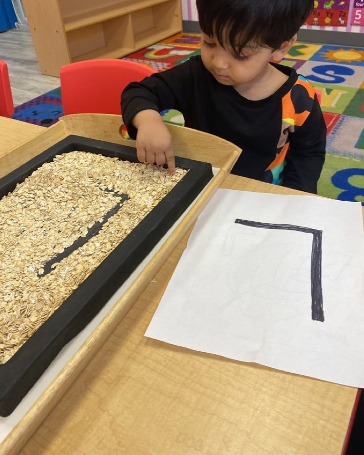 A young boy is playing with a tray of rice and a piece of paper with the letter l on it