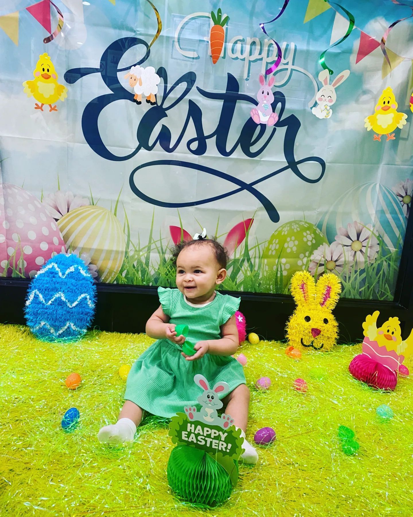 A baby in a green dress is sitting in a pile of easter eggs