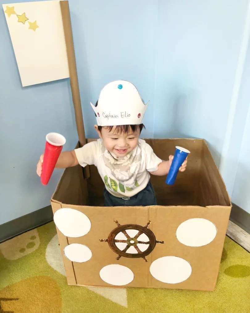 A little boy is playing in a cardboard box dressed as a pirate