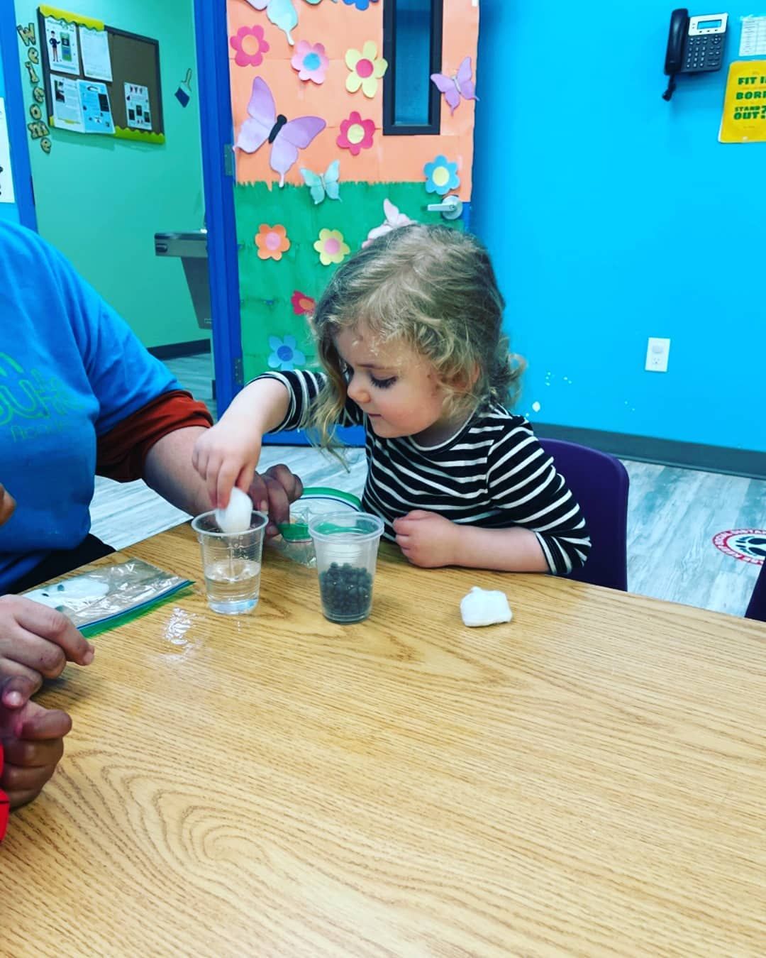 A little girl is sitting at a table playing with a glass of water