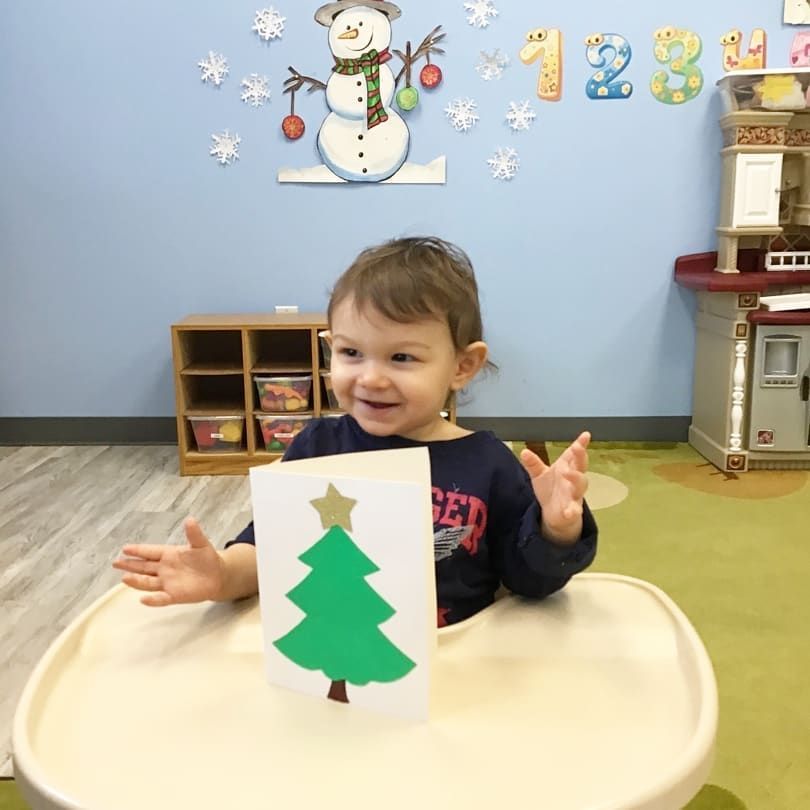 A little boy is sitting in a high chair holding a card with a christmas tree on it