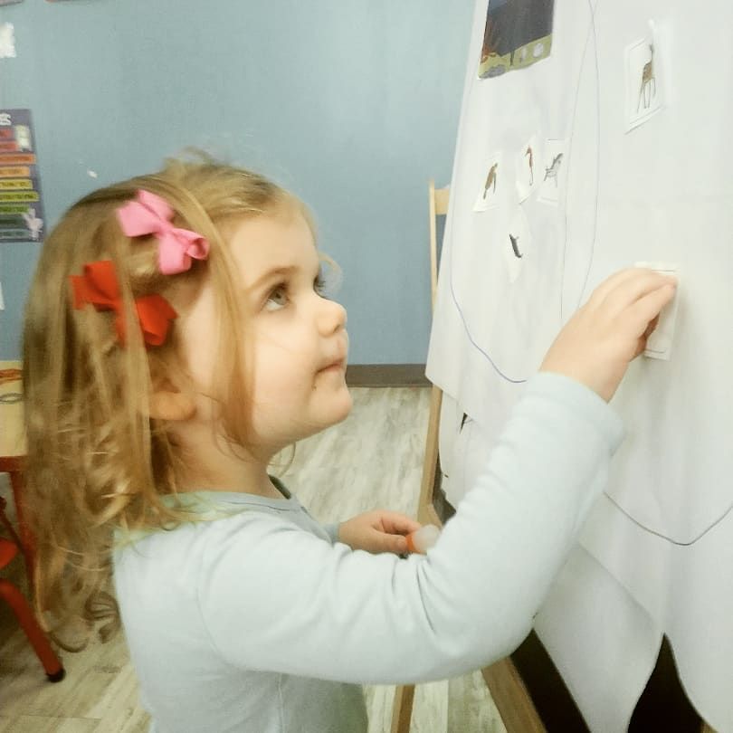 A little girl with a pink bow in her hair is drawing on a white board