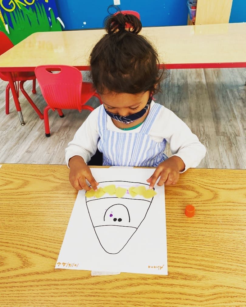 A little girl is sitting at a table with a piece of paper with a face on it