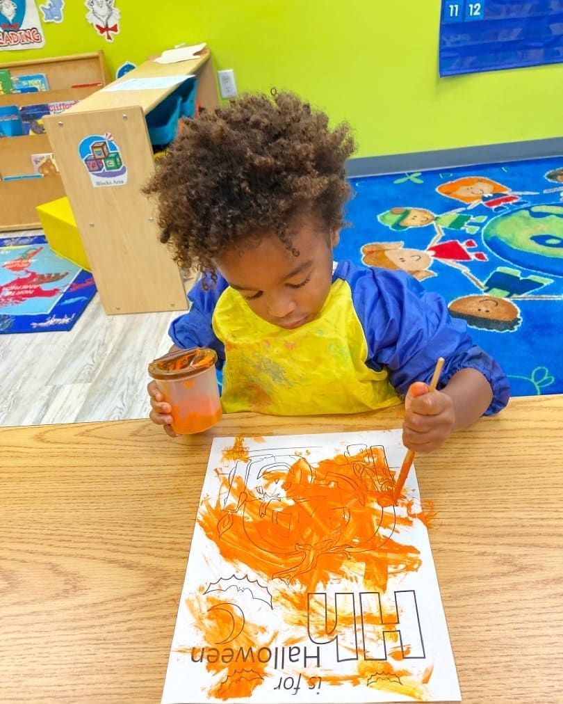 A young boy is sitting at a table painting with orange paint