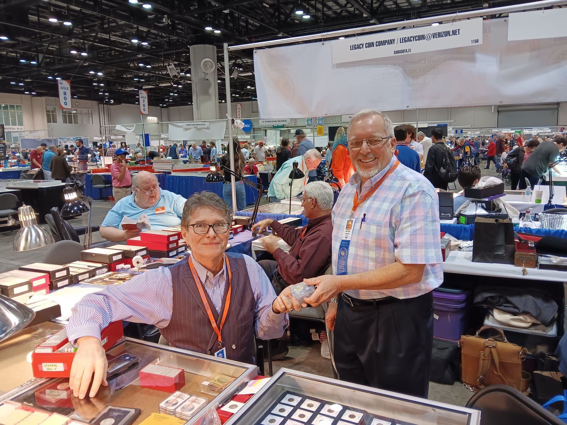 Two people, one handing a coin, at a coin show. Others browse the tables filled with coins.