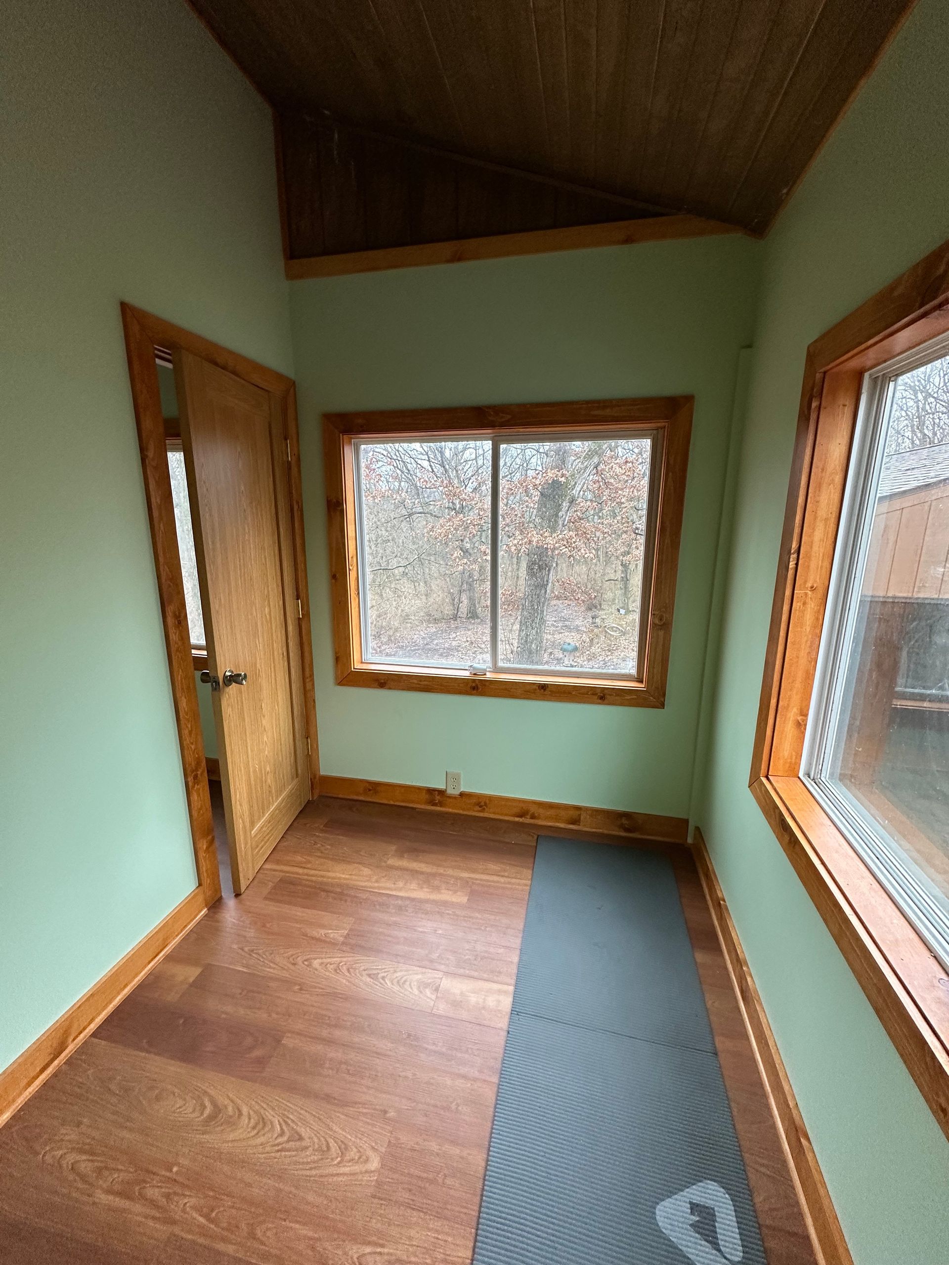 Small room with green walls, wood trim and door, two windows, yoga mat on the floor.
