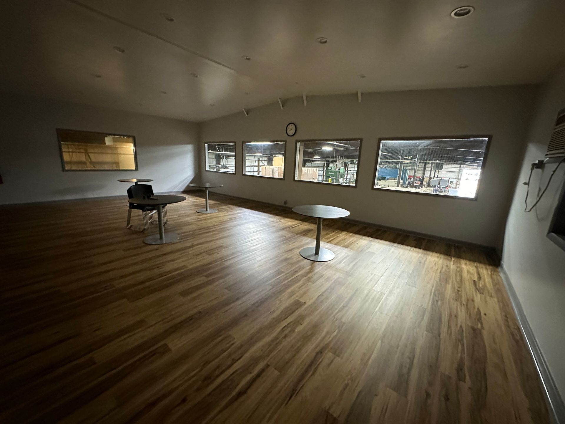 Empty room with wooden floors, small tables, and windows overlooking a factory.