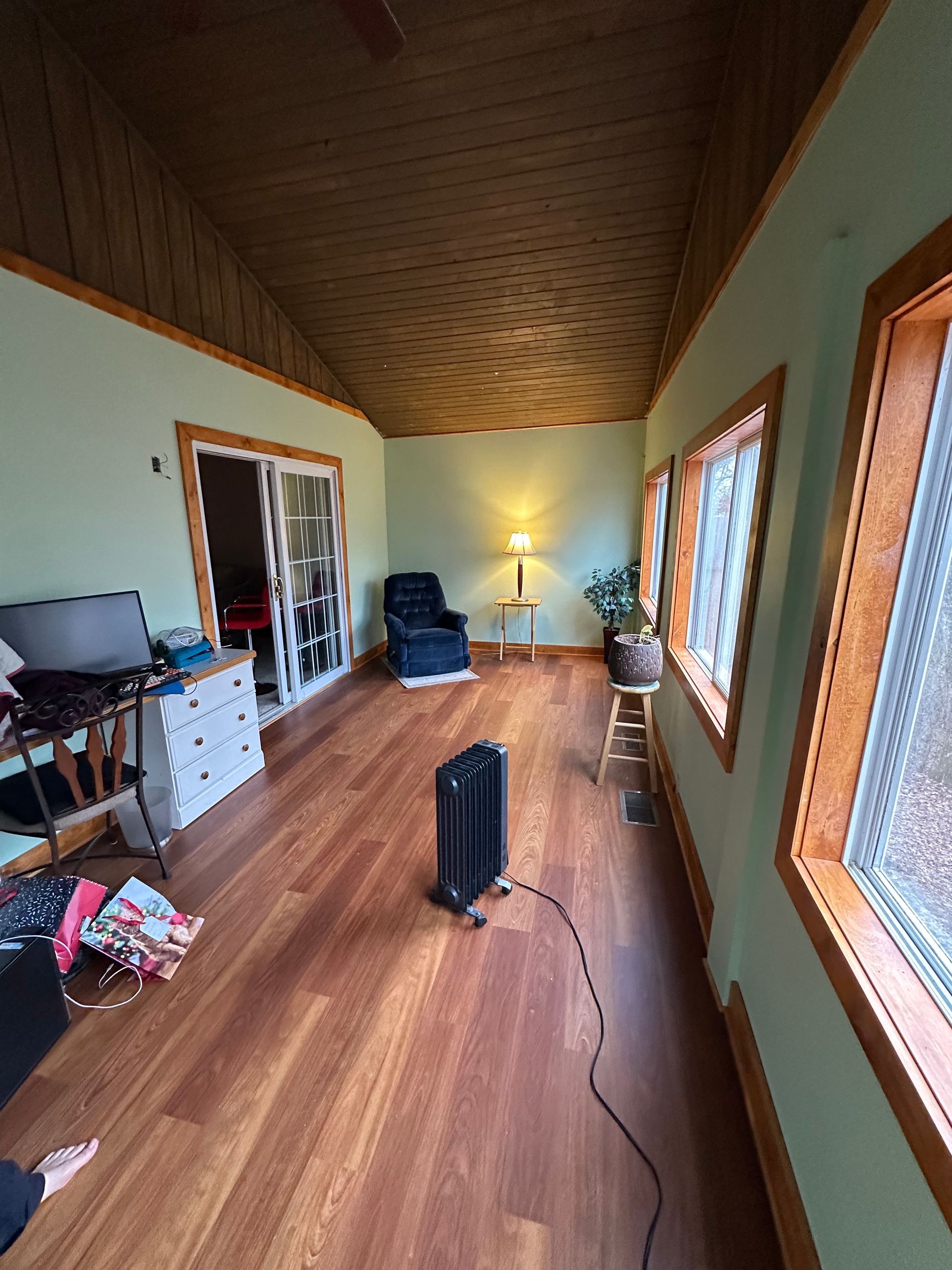 Room with wood floors, light green walls, wooden trim, windows, and a dark ceiling. Furnishings include a chair, dresser, and a heater.