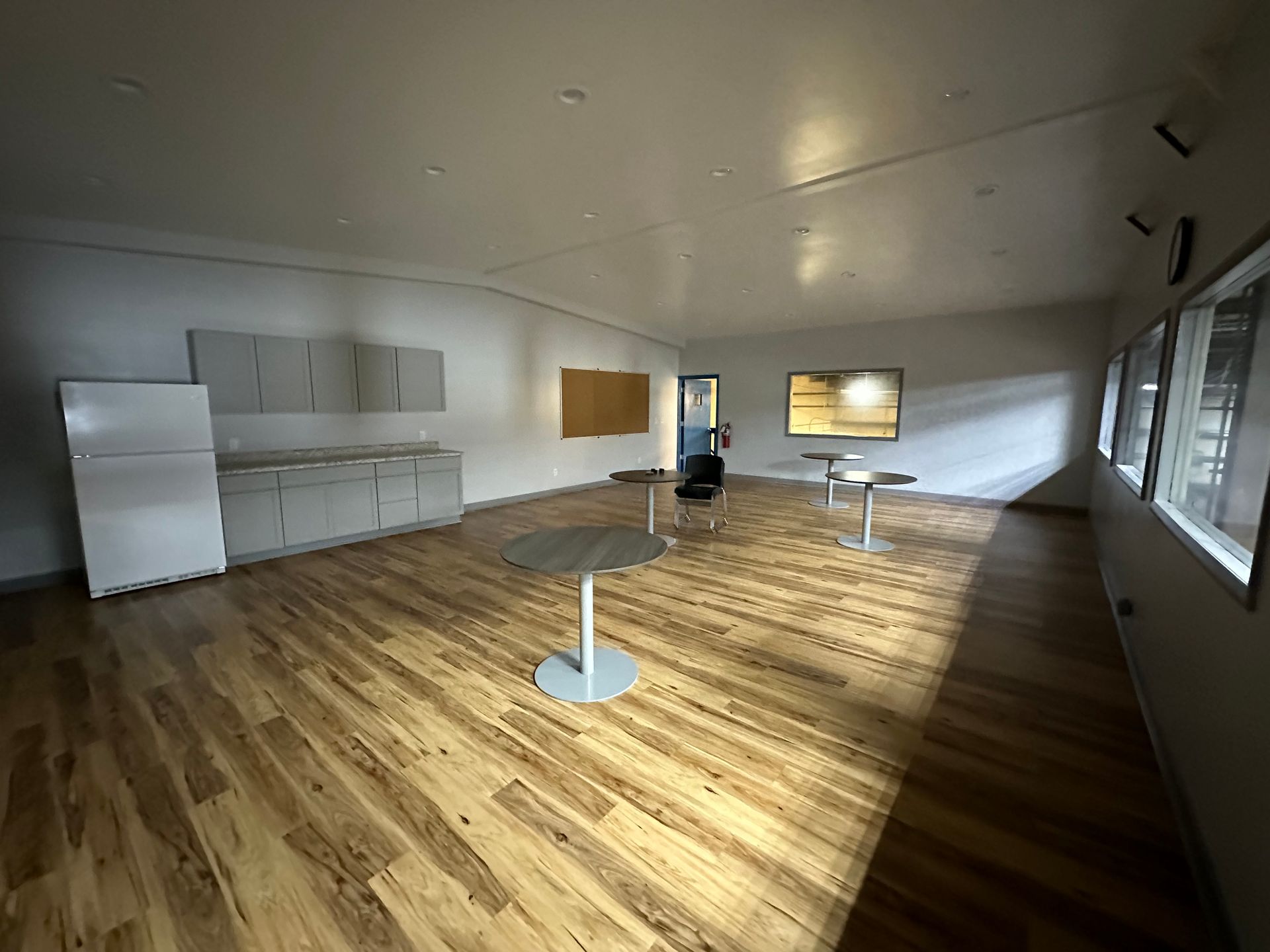 Empty room with wood floors, a refrigerator, cabinets, and small tables. Sunlight streams in from a window.