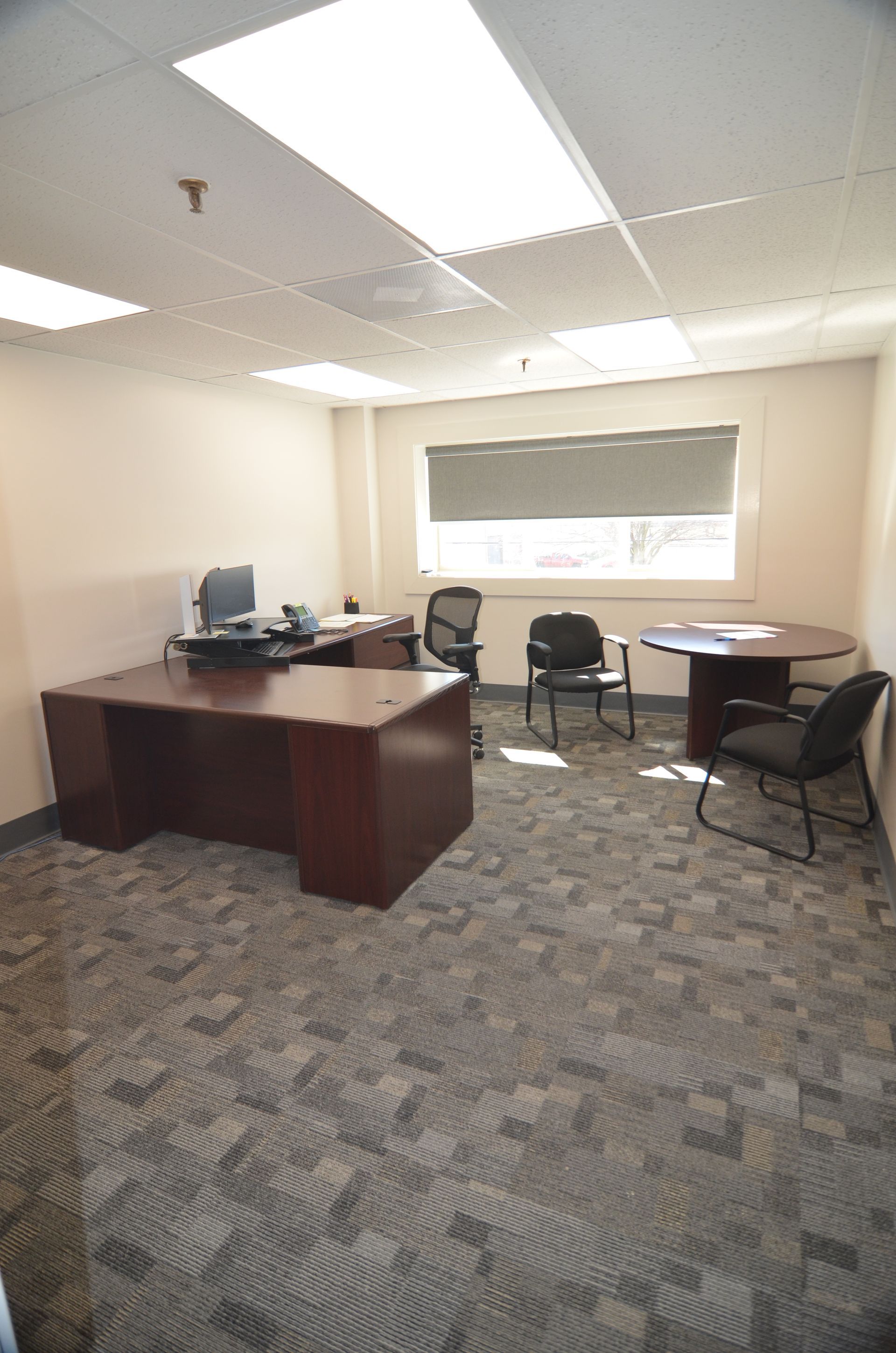 Office with desk, chairs, window, and patterned carpet. Beige walls, fluorescent lights.