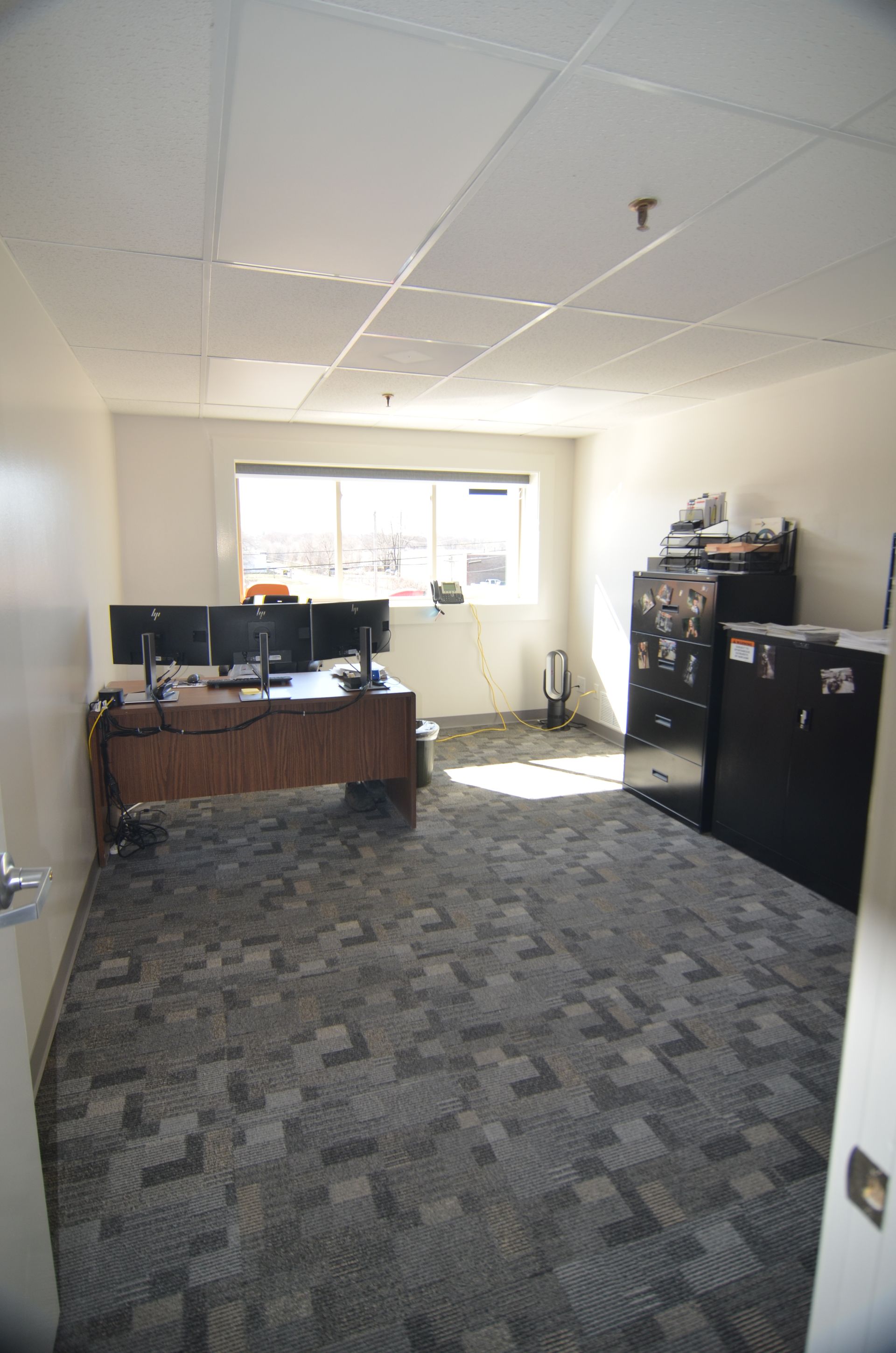 Empty office with desk, multiple monitors, and cabinets. Carpeted floor, window, and ceiling tiles.