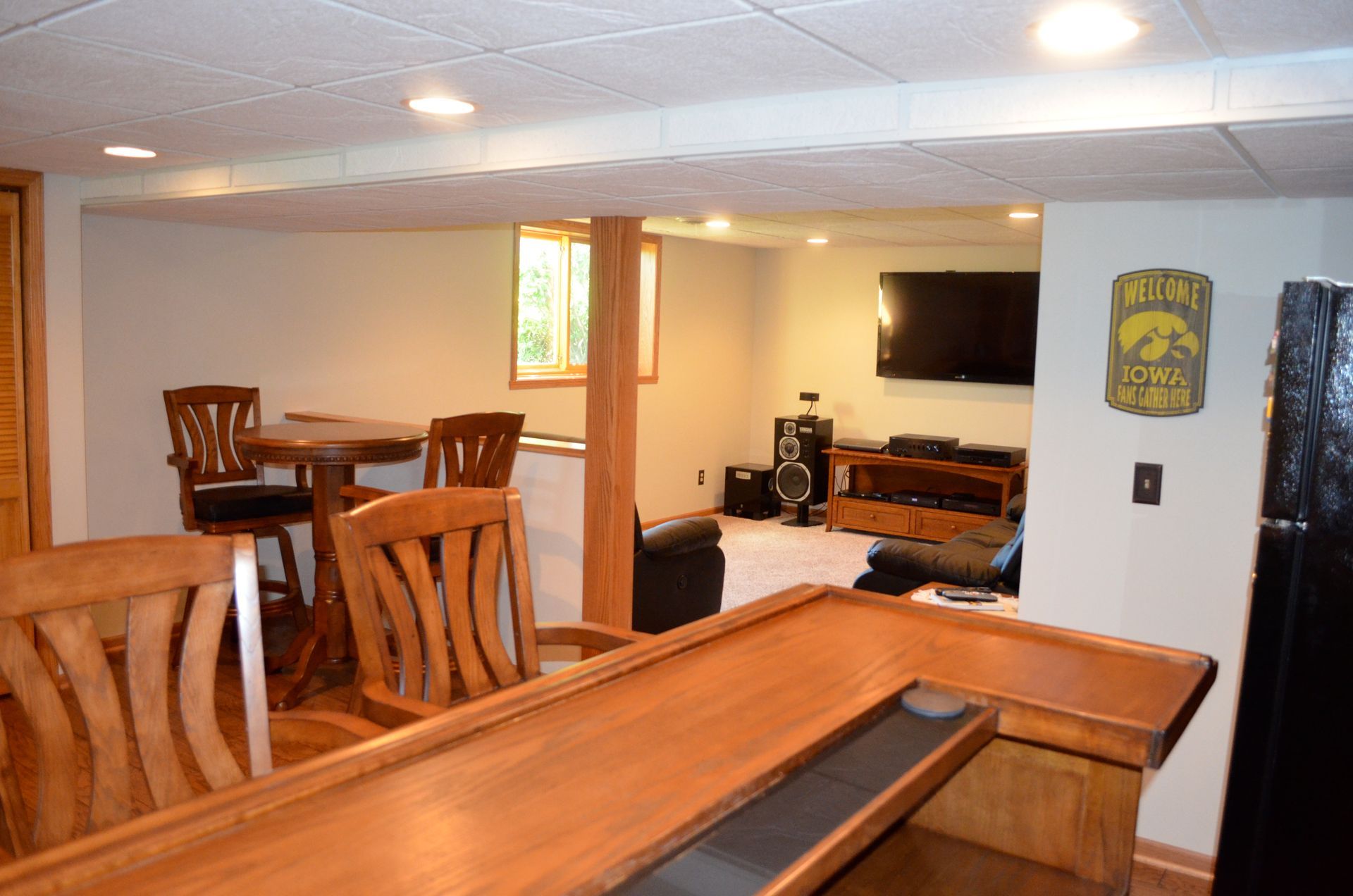 Basement bar area with wooden bar and stools, round table, TV, and Hawkeye sign.