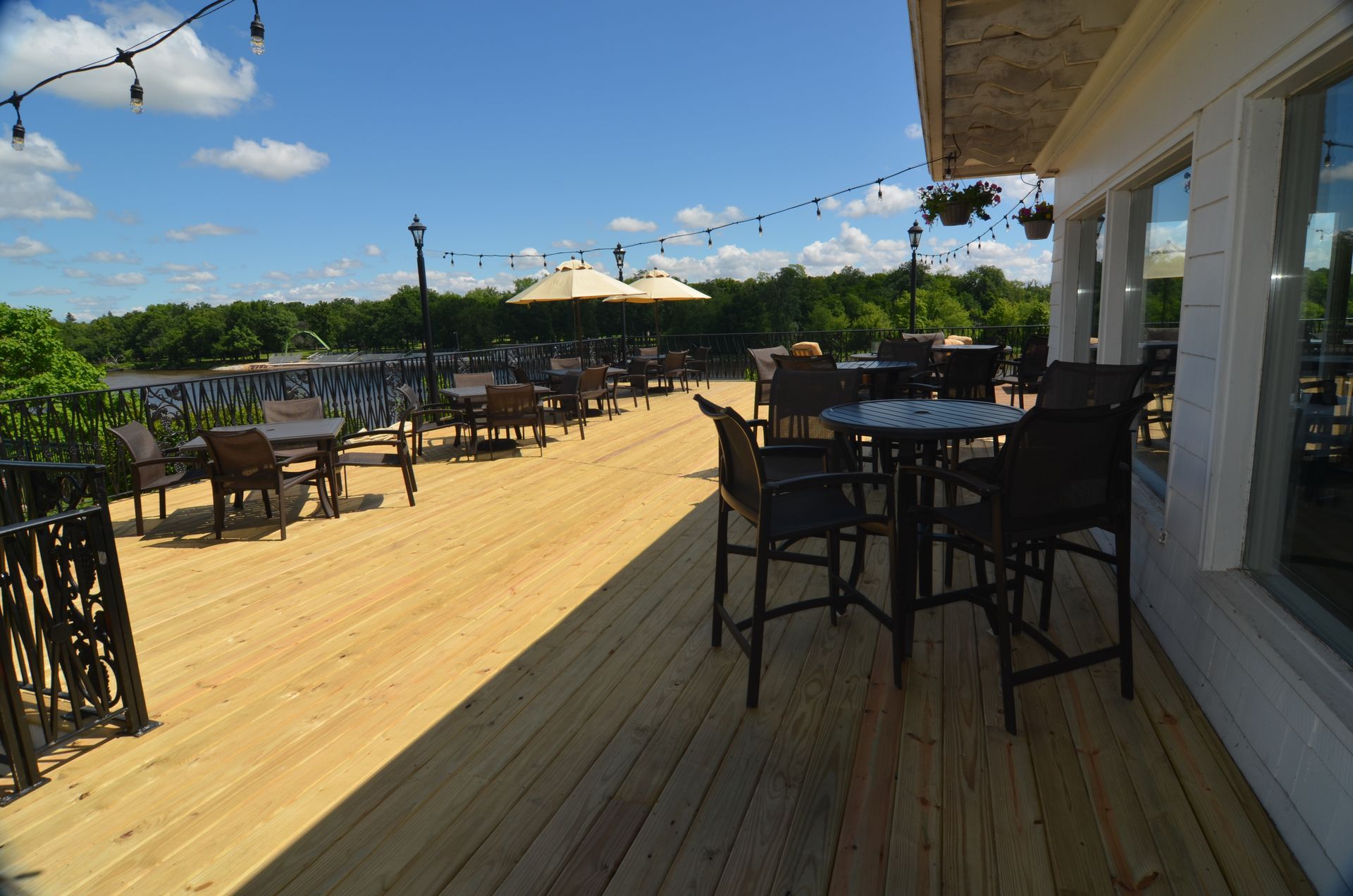 Outdoor restaurant patio with tables, chairs, and umbrellas overlooking a river under a blue sky.