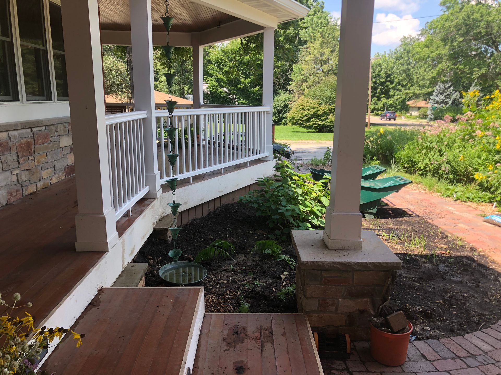 Porch with brick walkway and flowerbeds, white railing, and columns. Green trees in the background.