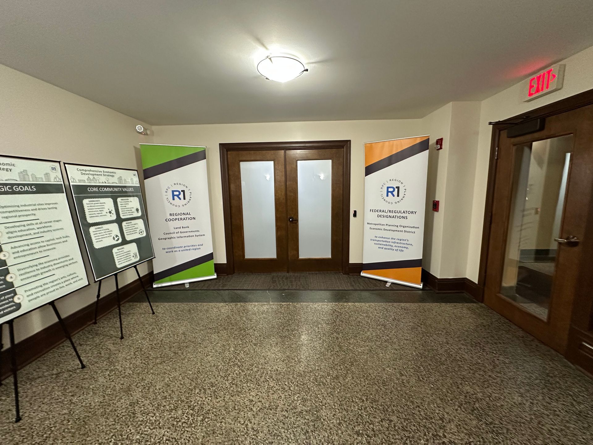 Hallway with posters and banners: doors in the center, exit sign to the right, and display boards to the left.