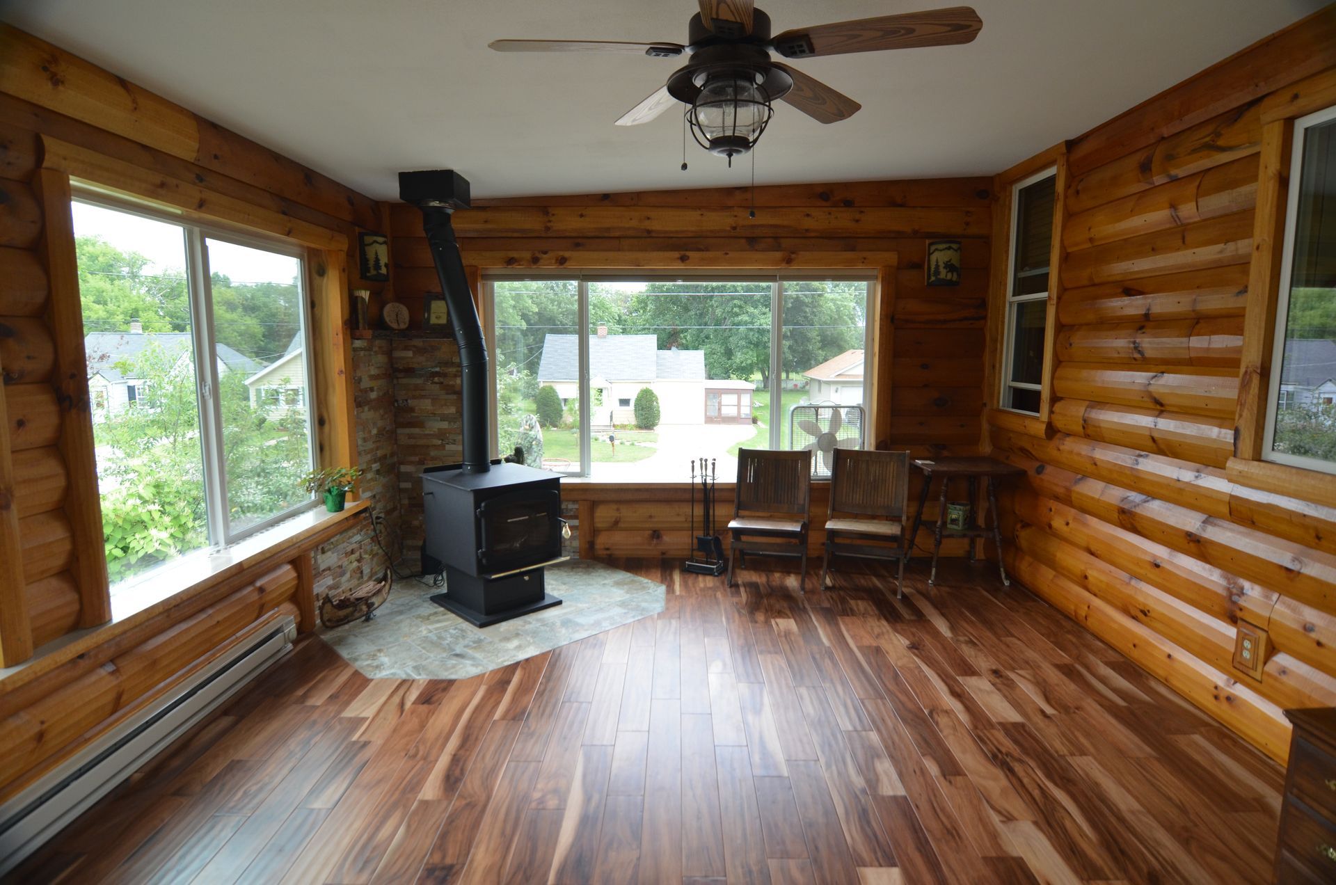 Sunroom with wood-paneled walls, large windows overlooking a yard, and a wood-burning stove on a stone hearth.