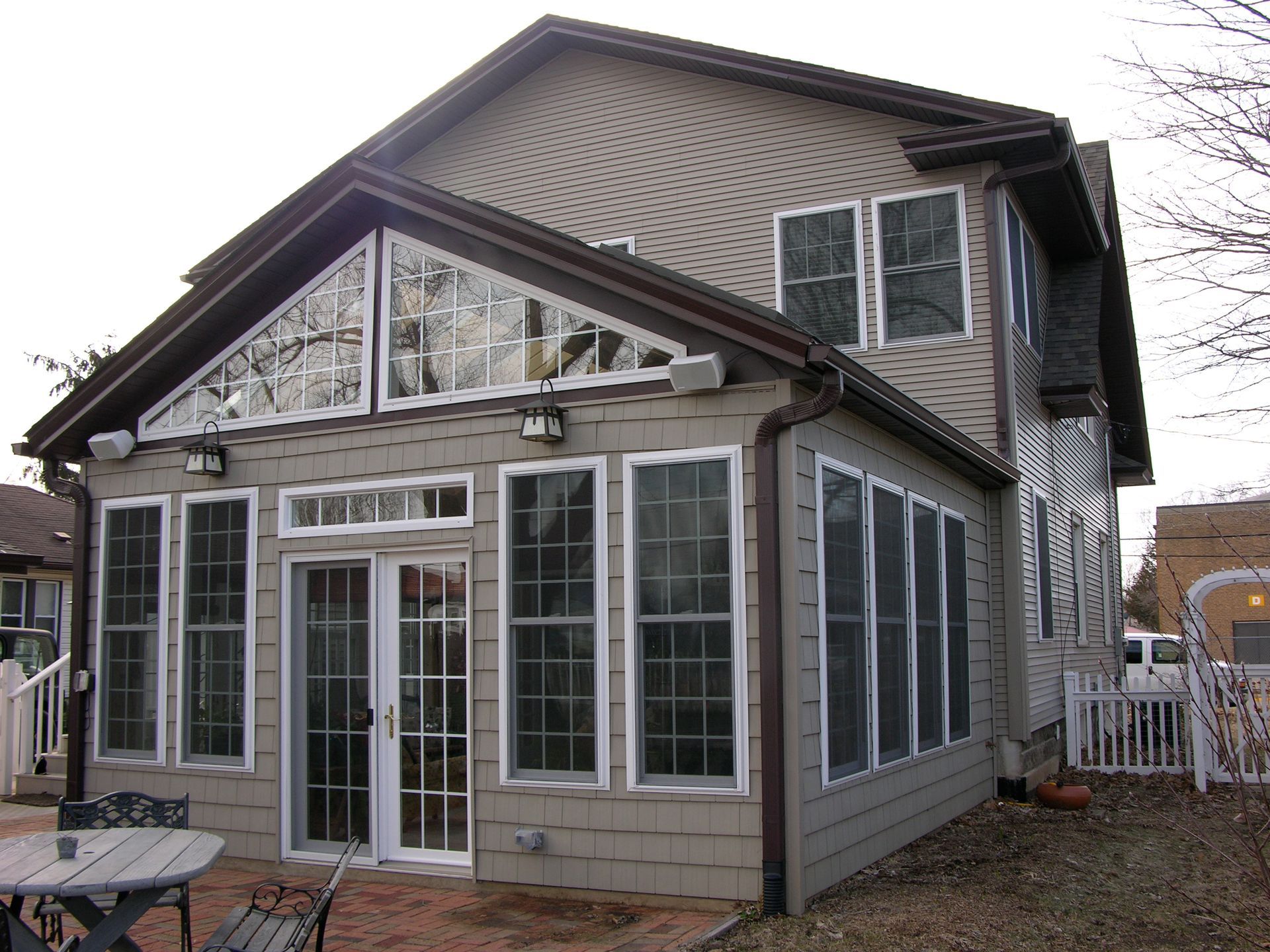 Tan-sided house with a sunroom addition, featuring large windows and glass doors, brown trim, and a brick patio.