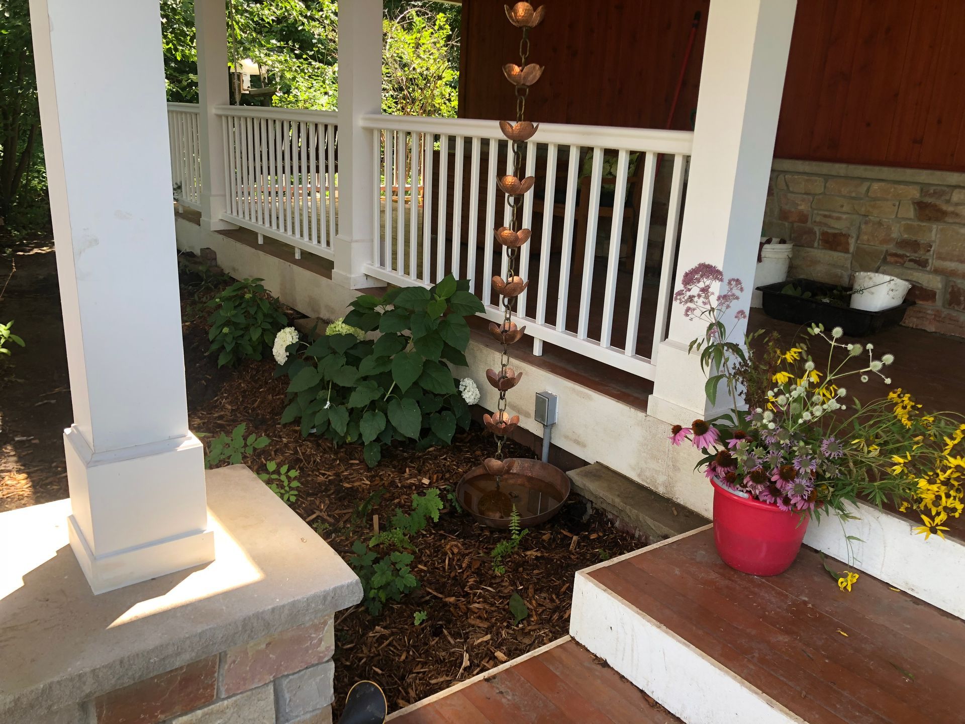 Porch with white railing, stone columns, garden bed with plants, copper rain chain, and flower pot.