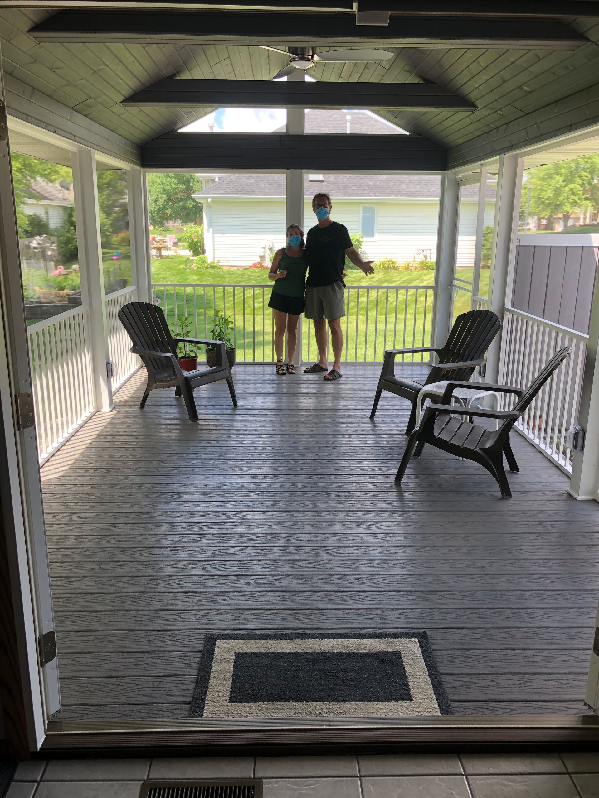 Two people wearing masks on a screened porch. Gray deck, chairs, and welcome mat. Backyard in background.