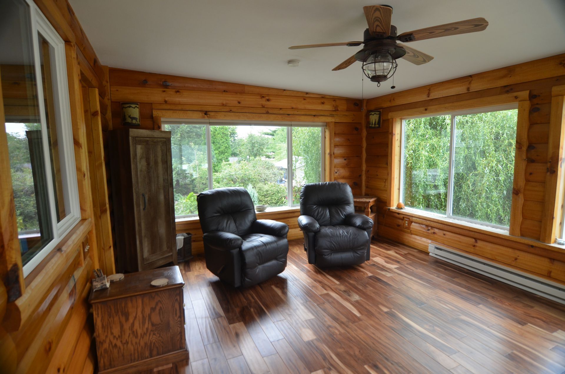 Sunroom with wood walls, two black recliners, and large windows overlooking trees.