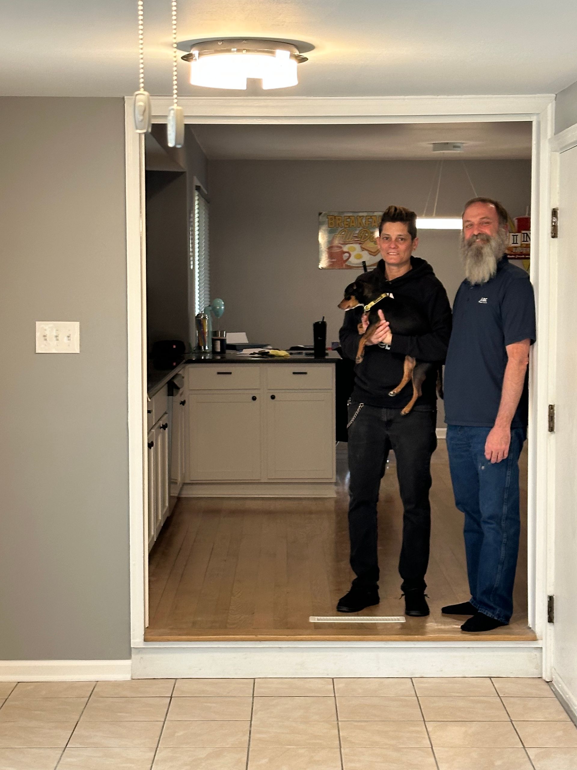 Two people, one holding a small dog, stand in a doorway; kitchen in the background.